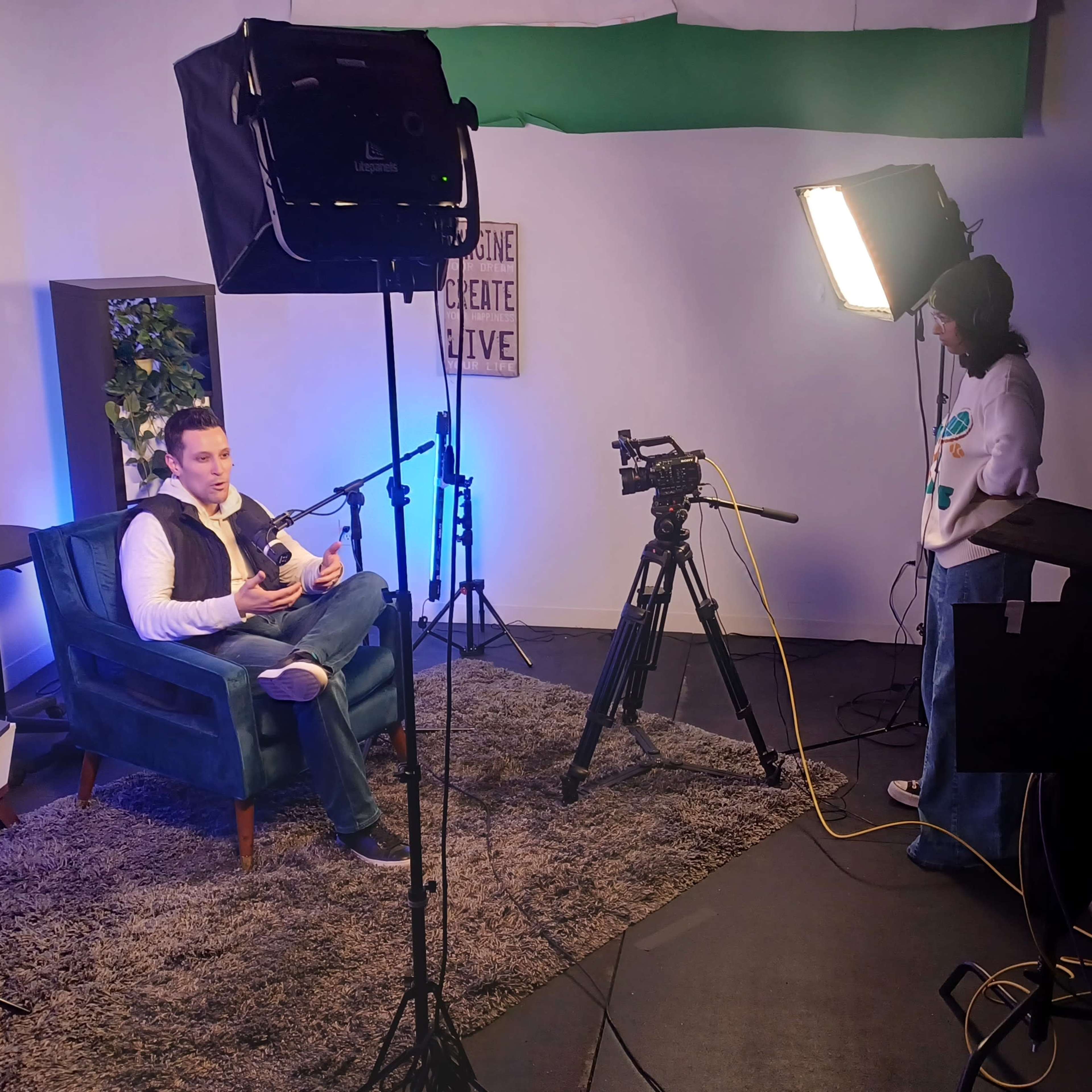 A man sits in a blue chair while a person stands beside a camera in a well-lit studio setup.