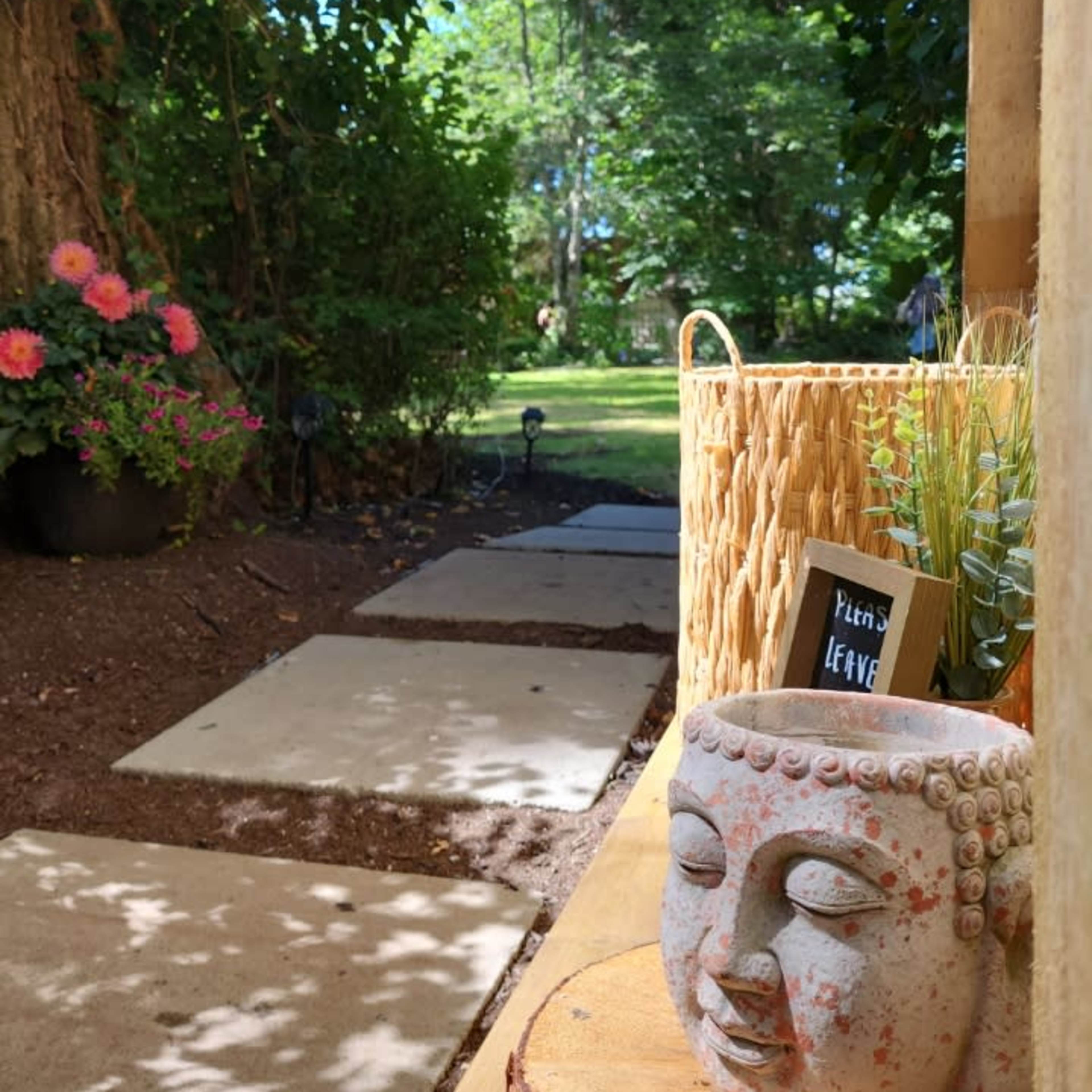 A stone Buddha head rests on a wooden slab next to a basket with greenery, framed by a pathway and blooming flowers in a garden setting.