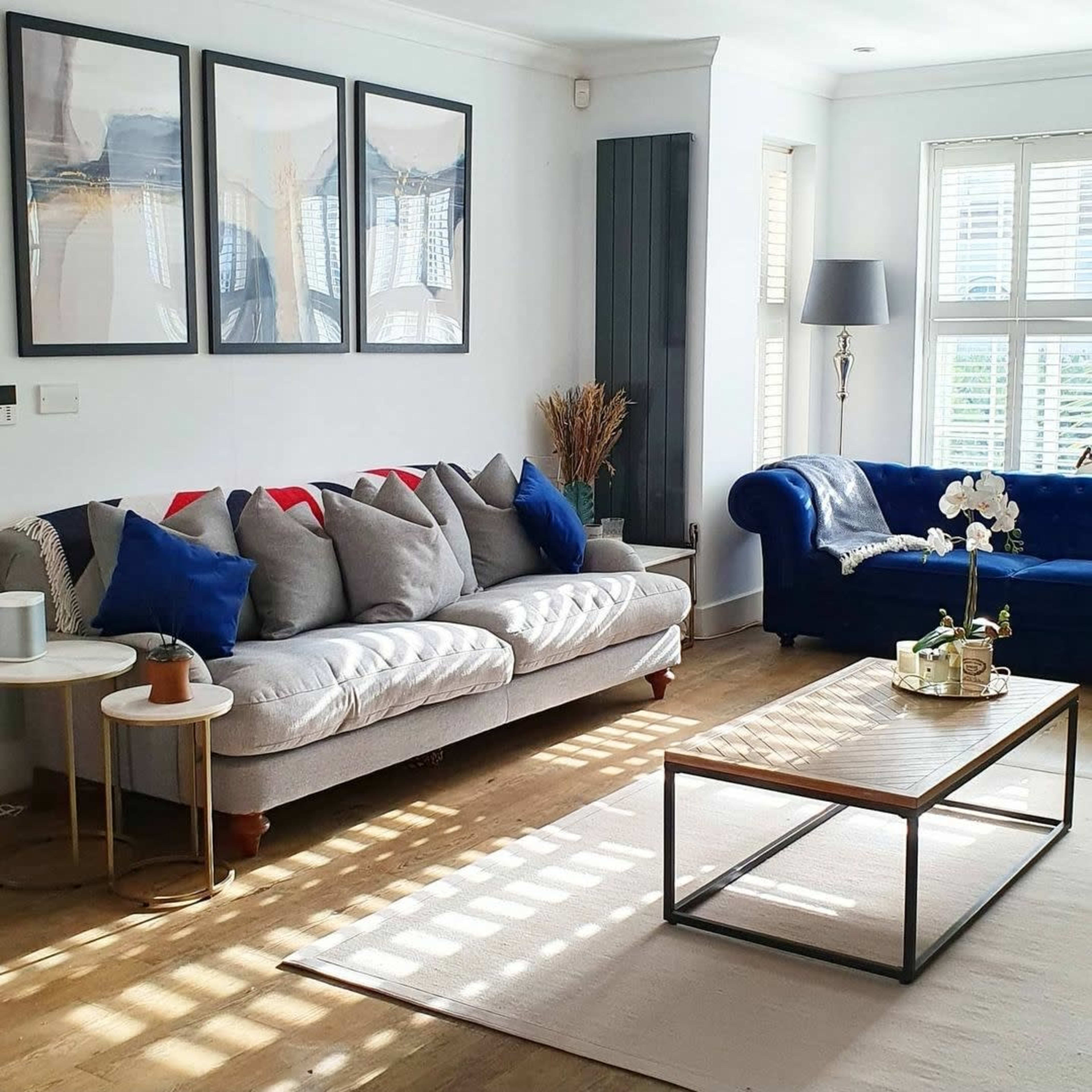 A living room features a gray sofa with colorful cushions, a blue chair, a wooden coffee table, and framed artwork on the walls.