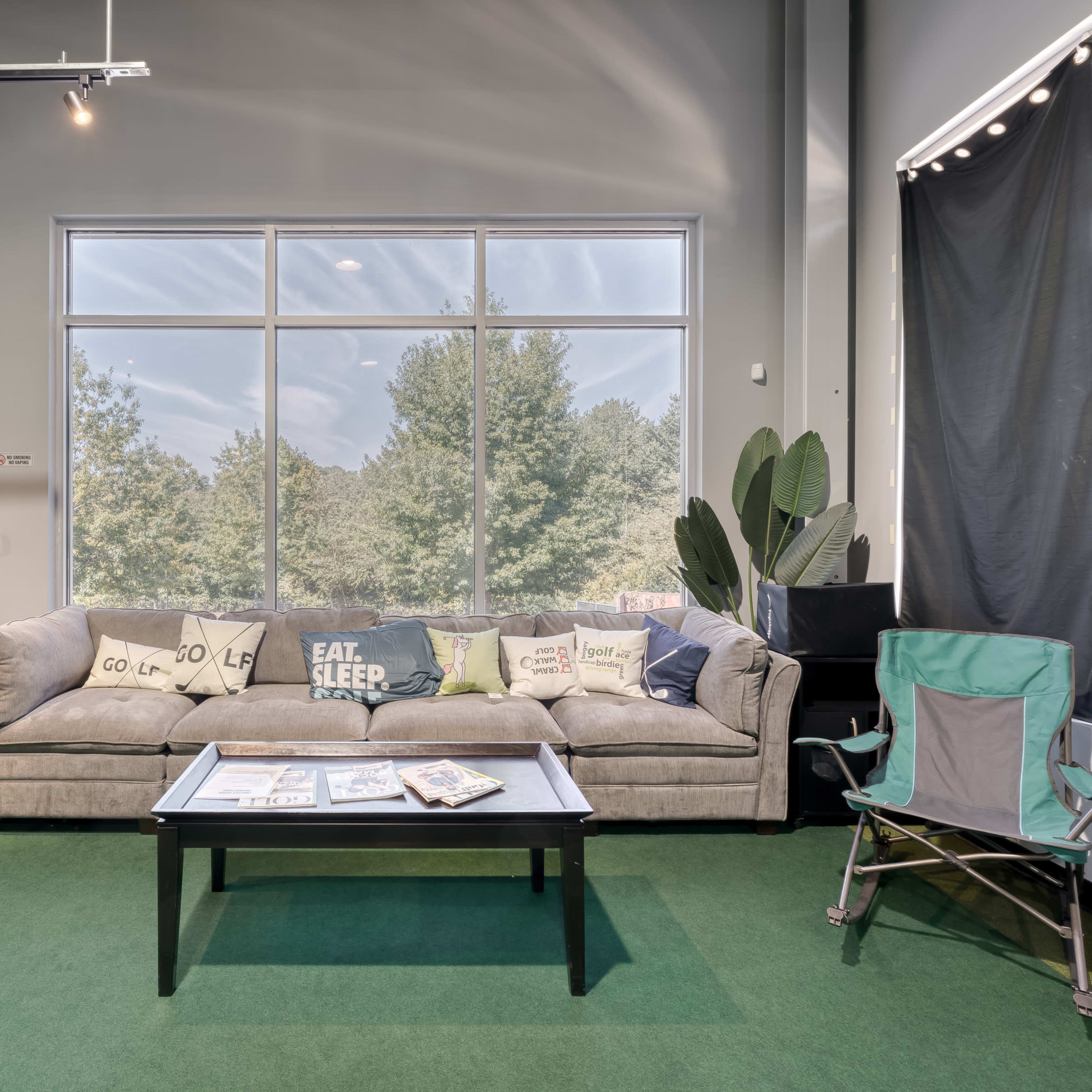 A cozy lounge area featuring a gray sofa with decorative pillows, a black coffee table, a green folding chair, and large windows overlooking trees.