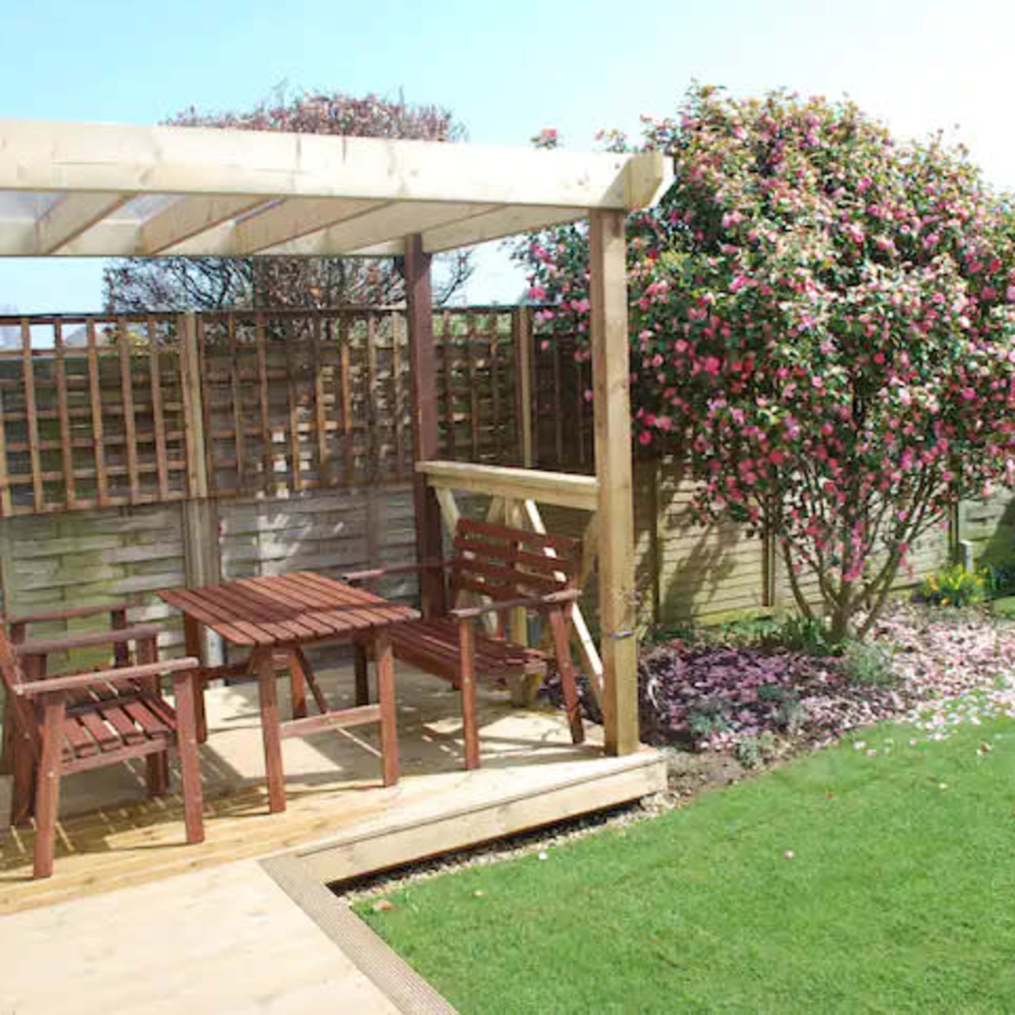 A wooden patio with a table and chairs is situated next to a flowering bush in a well-maintained garden.