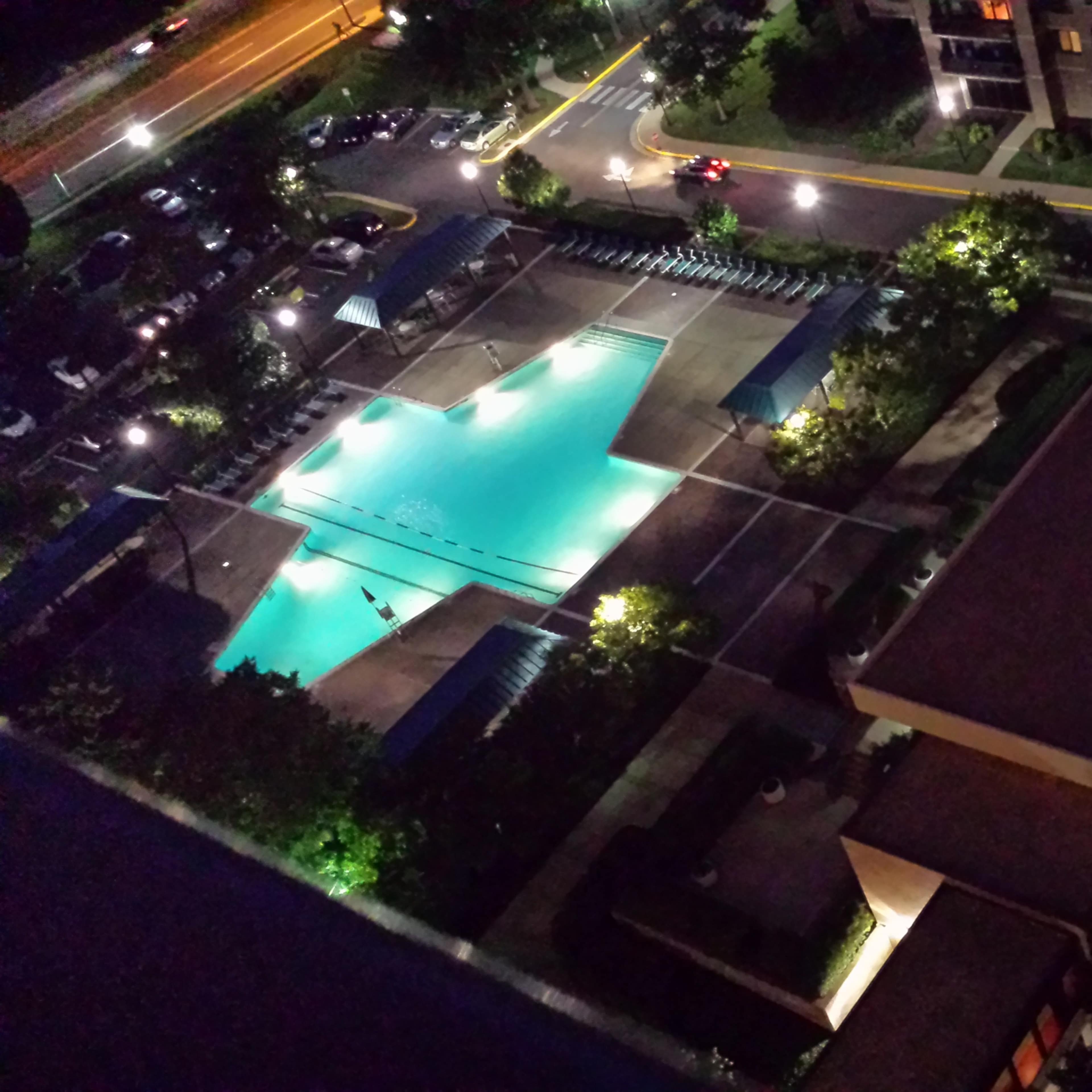 An aerial view of a illuminated swimming pool area surrounded by buildings and parked cars at night.