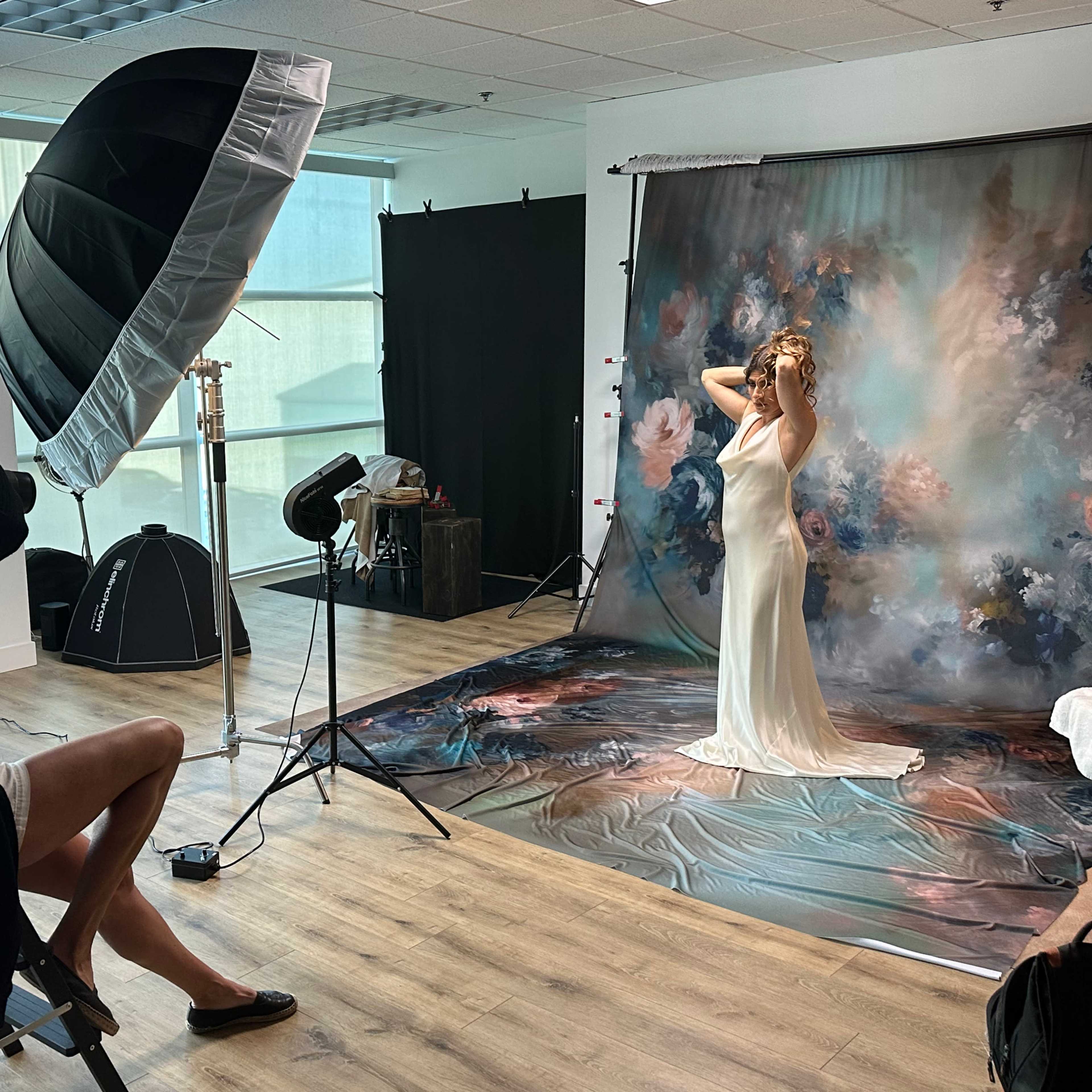 A photographer captures a model in a flowing dress posed in front of a painted backdrop in a studio setting.