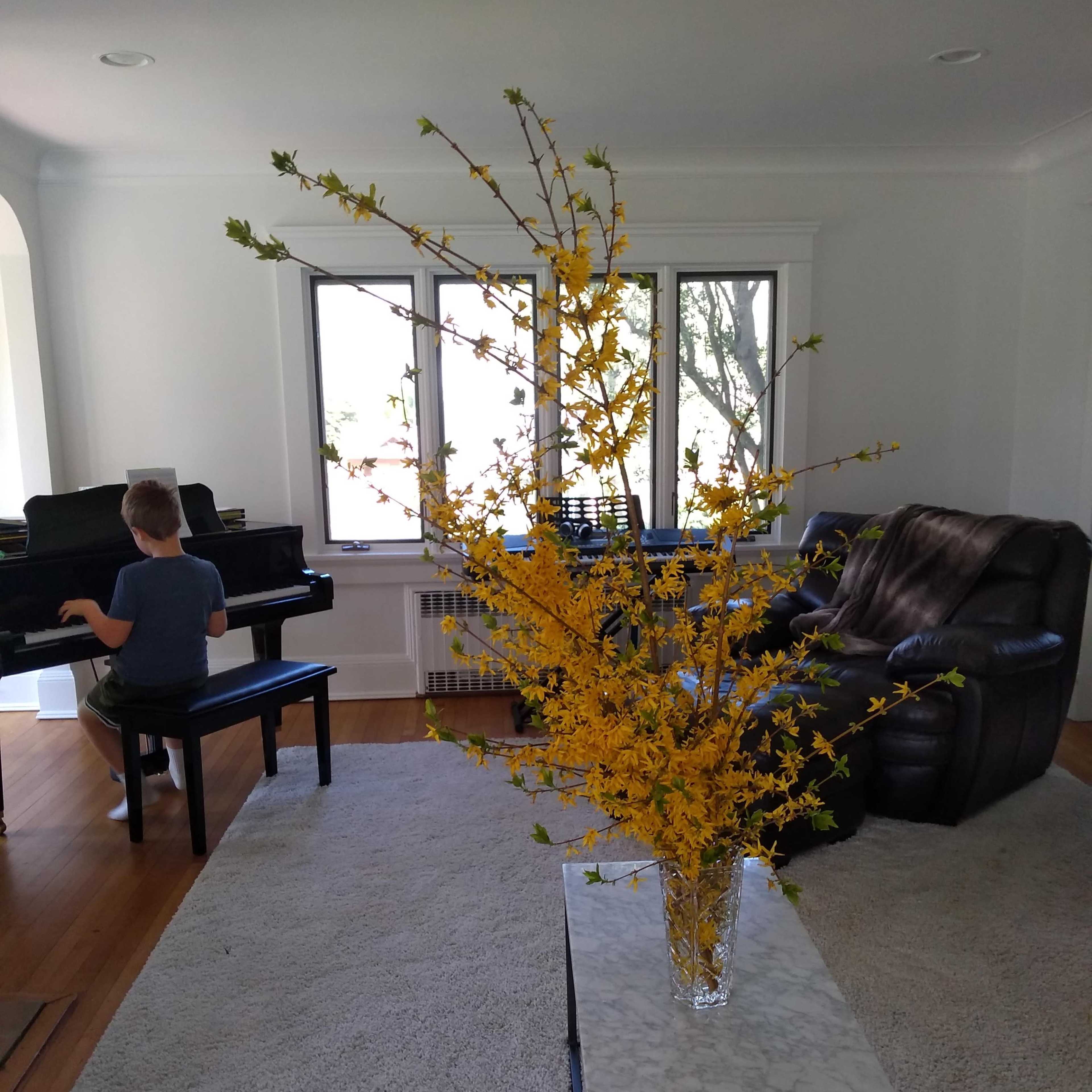 A child is playing a piano in a living room with a large vase of yellow flowers on a coffee table.