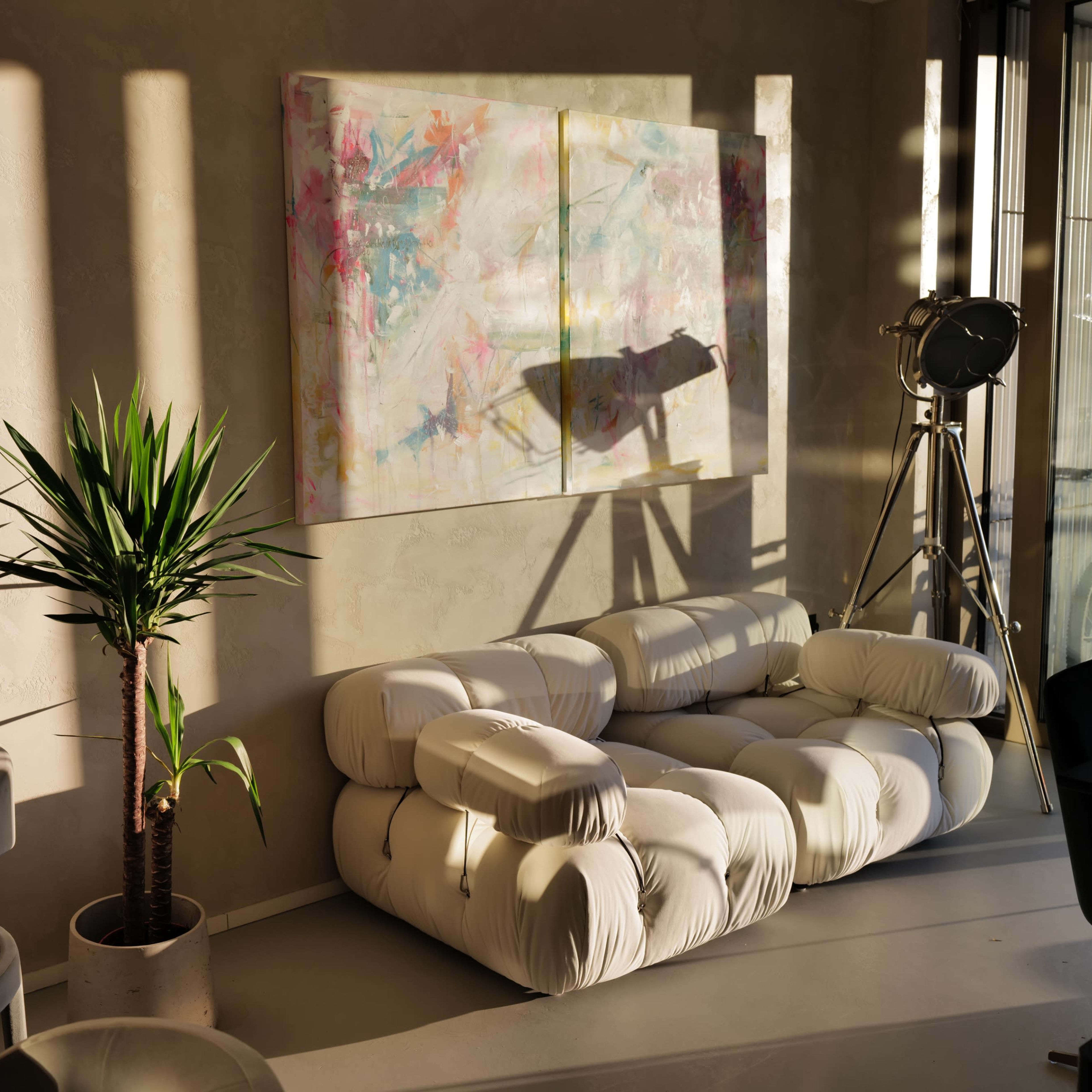 A modern living space featuring a white sofa, a round table, a potted plant, and a colorful abstract painting on the wall, with sunlight casting shadows across the room.