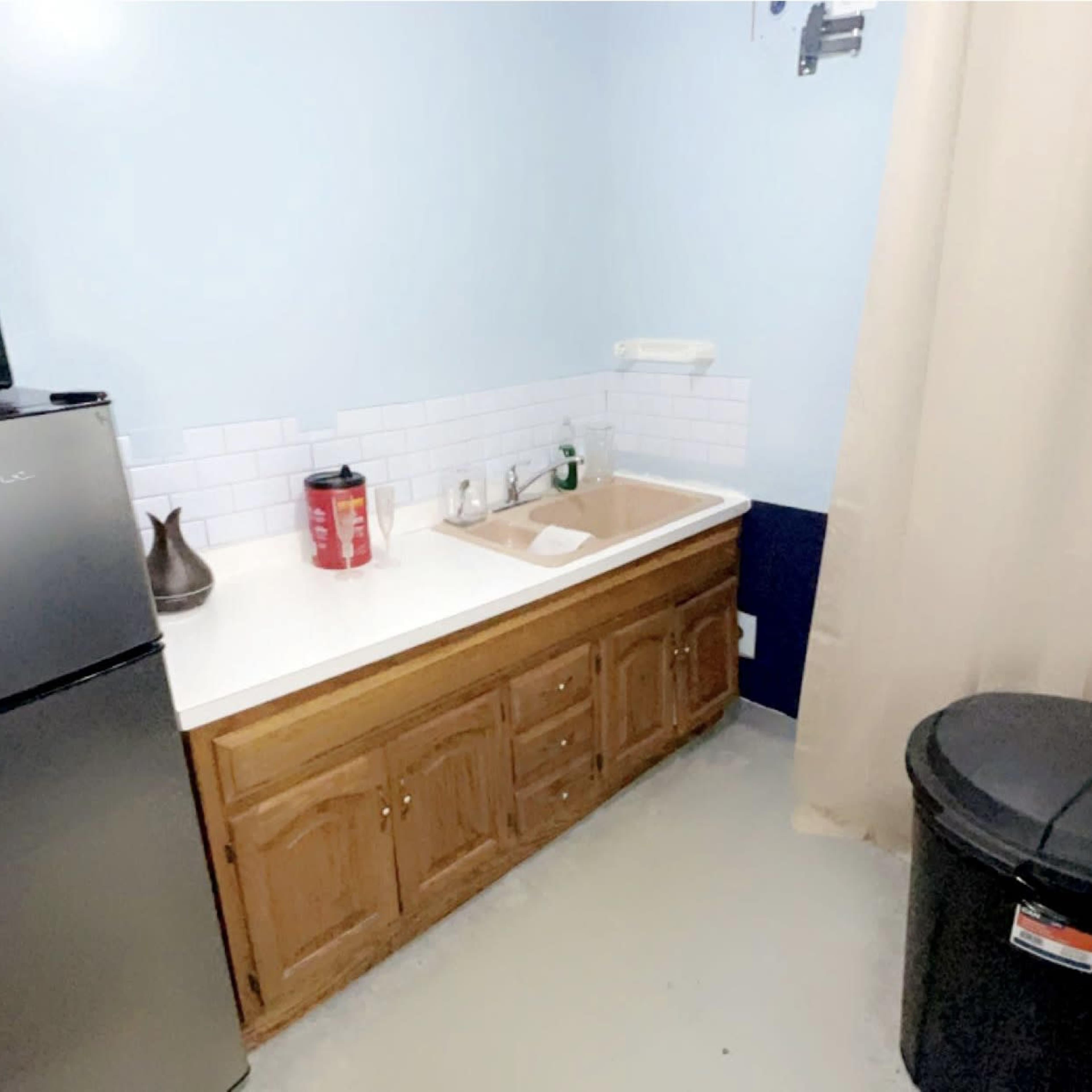 A small kitchen area with a stainless steel refrigerator, a sink counter with a faucet, and a black trash can beside it.
