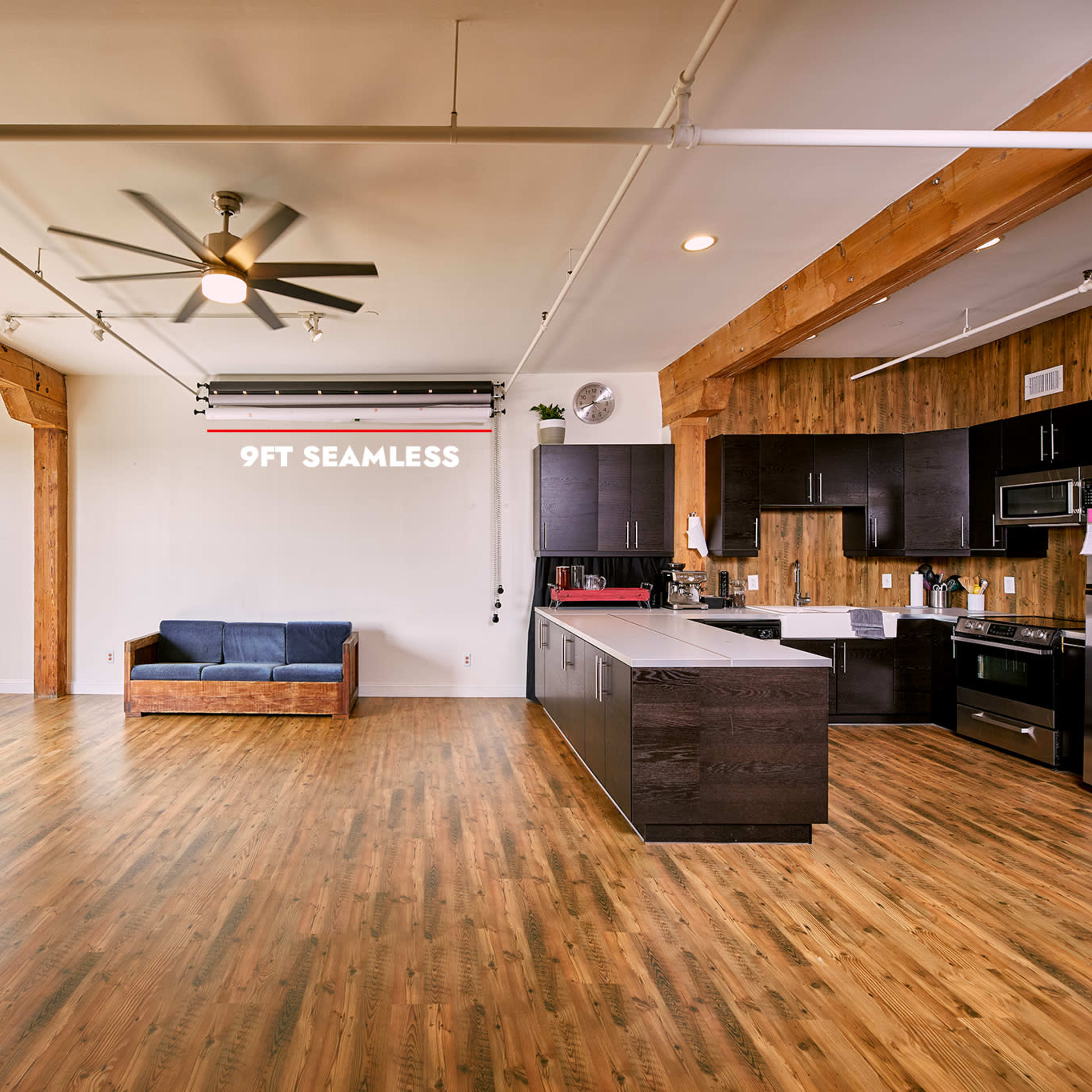An open living space features a kitchen with dark cabinets, a wooden accent wall, a blue couch, and large windows allowing natural light to fill the room.