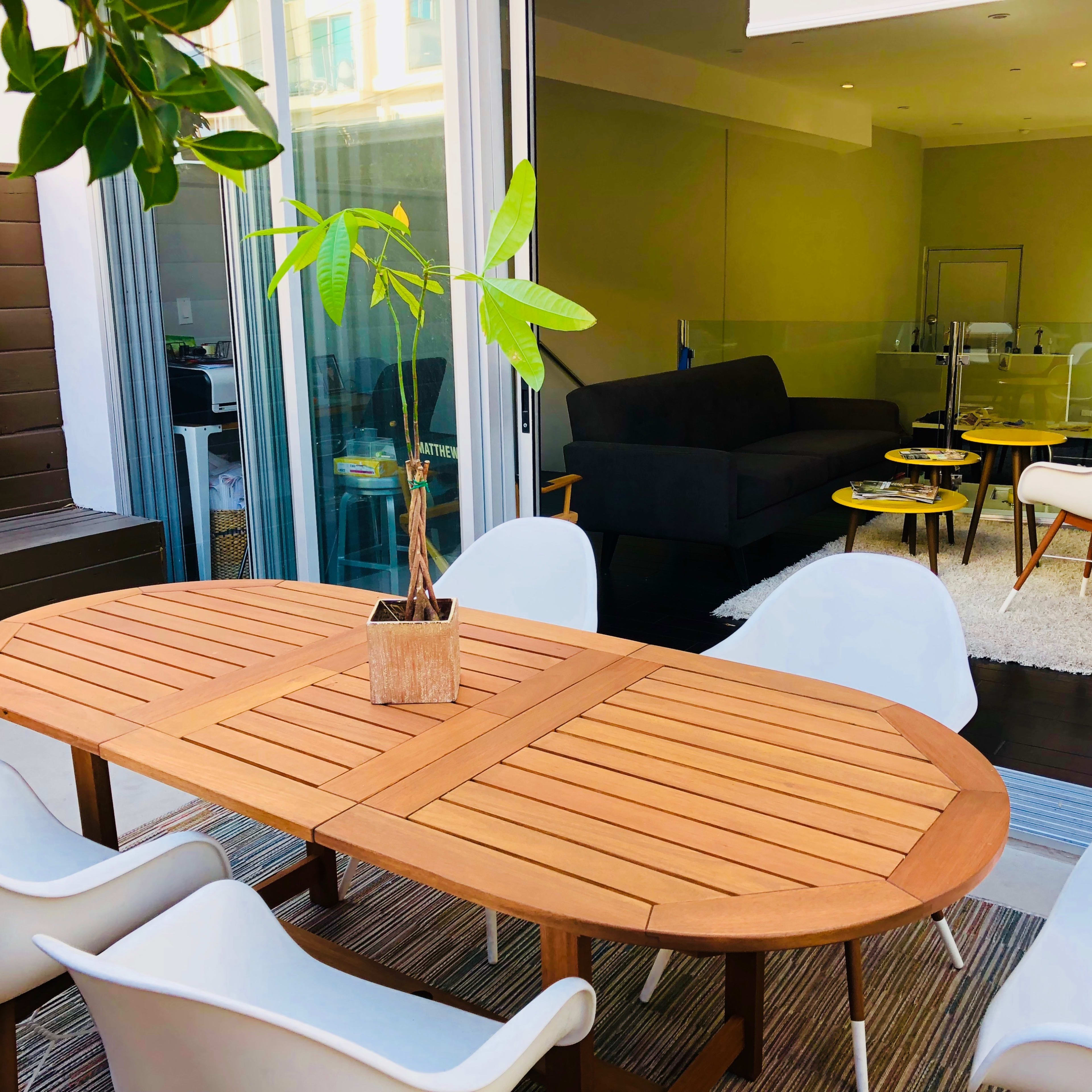 A wooden oval dining table with a potted plant in the center is surrounded by white chairs on a patio, leading into a brightly lit indoor space with a couch and two small tables.