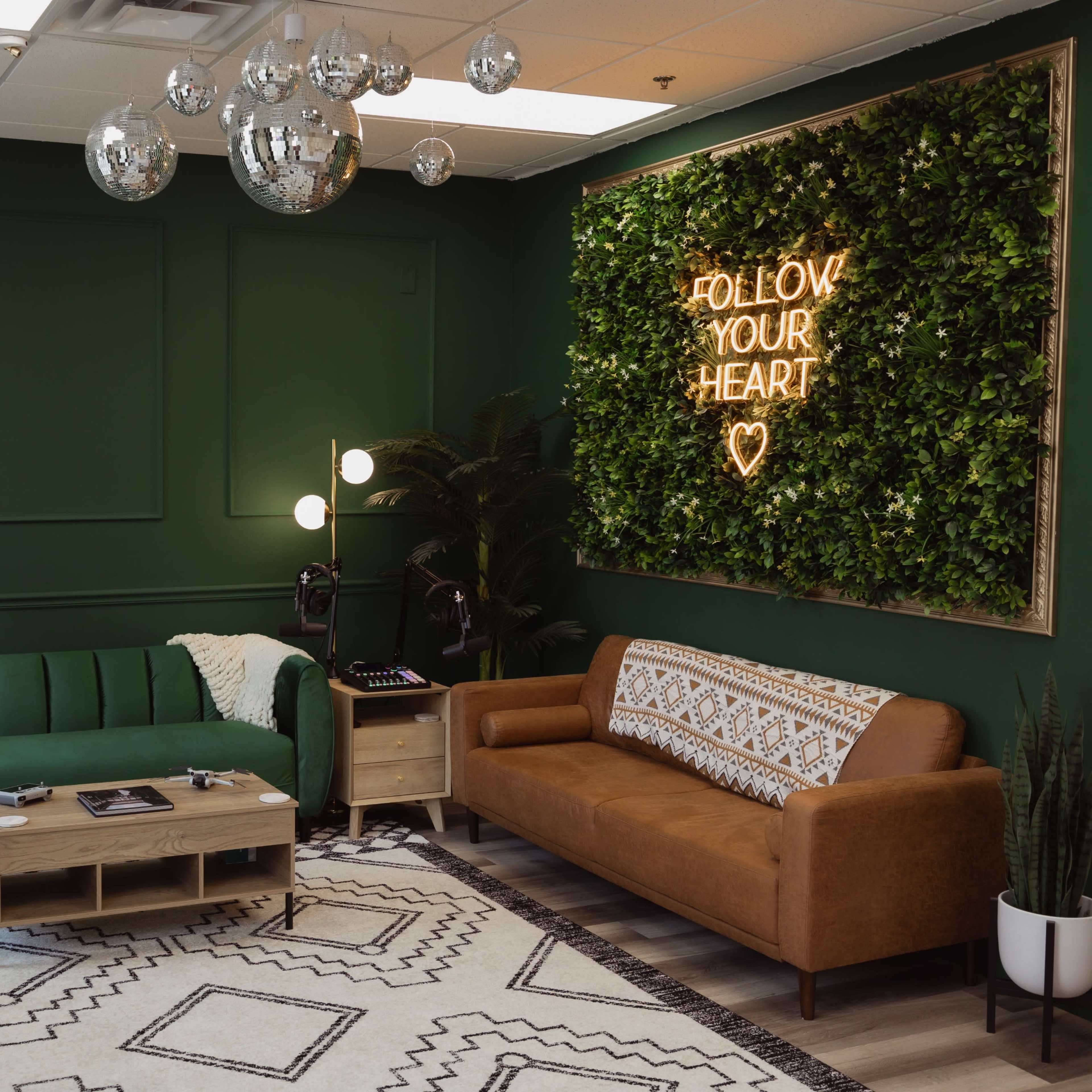 A modern lounge area featuring a green wall, two sofas, a hanging chair, a wooden coffee table, and a neon sign surrounded by plants.