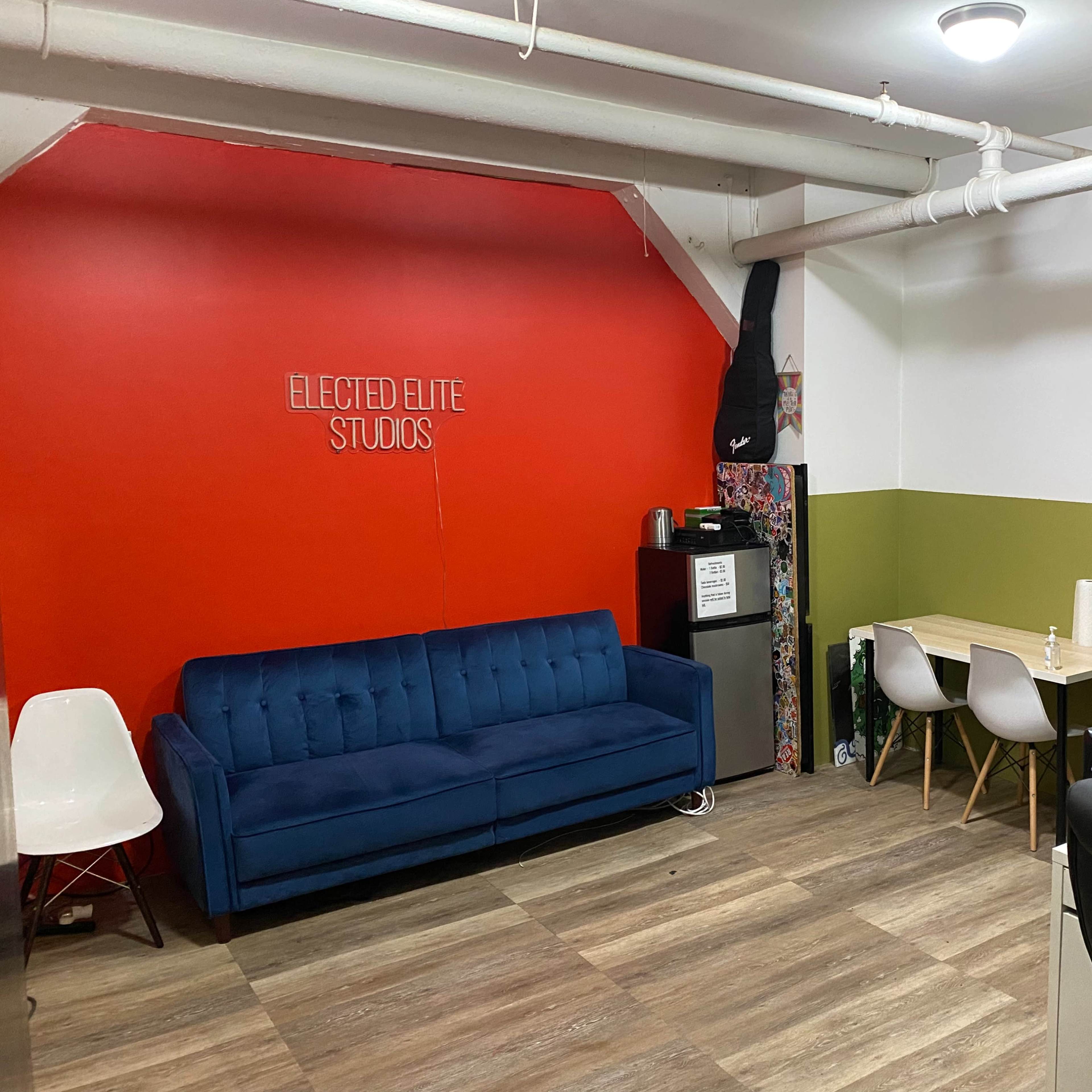 An interior studio with a red accent wall, a blue couch, a mini-fridge, a desk with equipment, and white chairs.