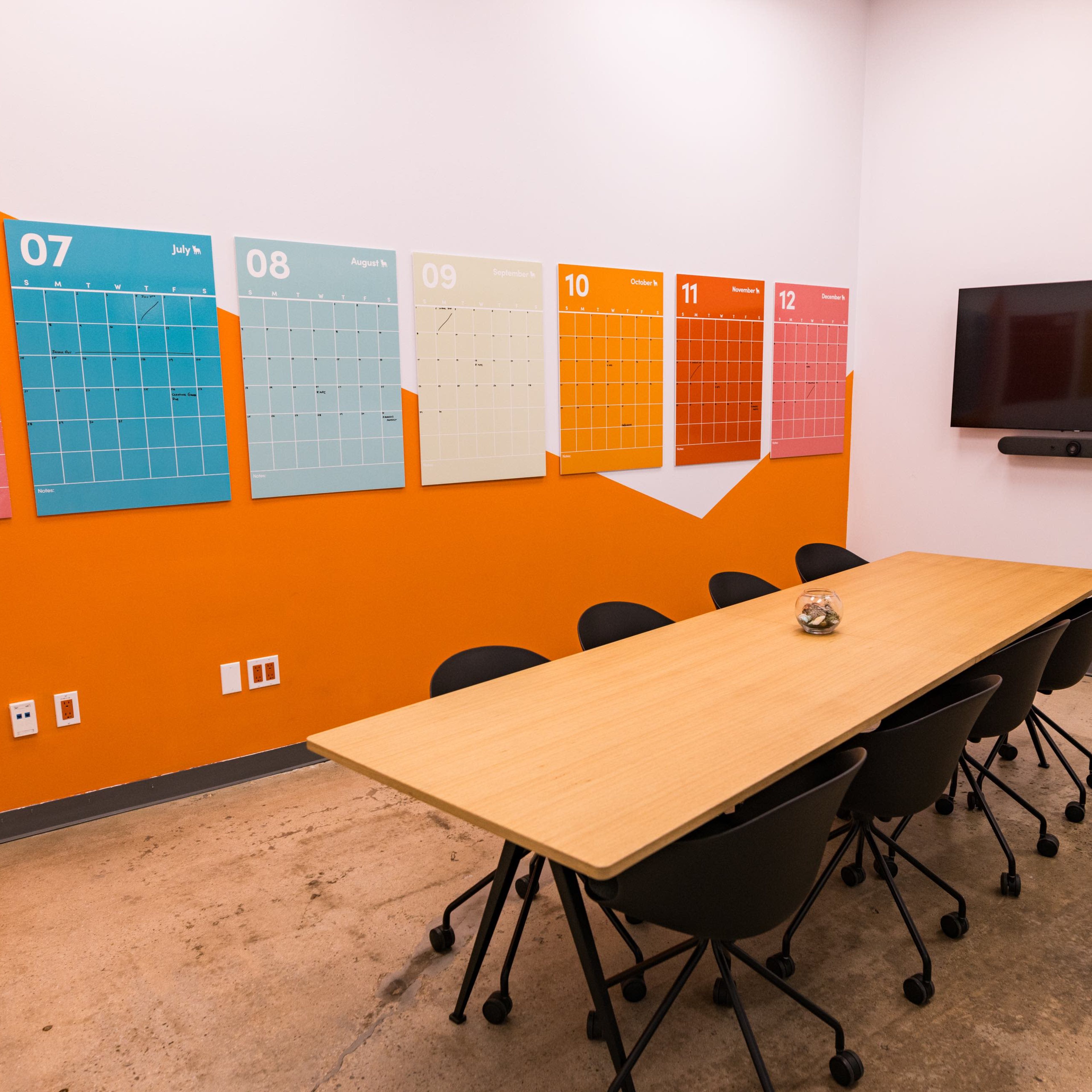 A meeting room features a long wooden table surrounded by black chairs, with colorful monthly calendars on the wall and a television mounted nearby.