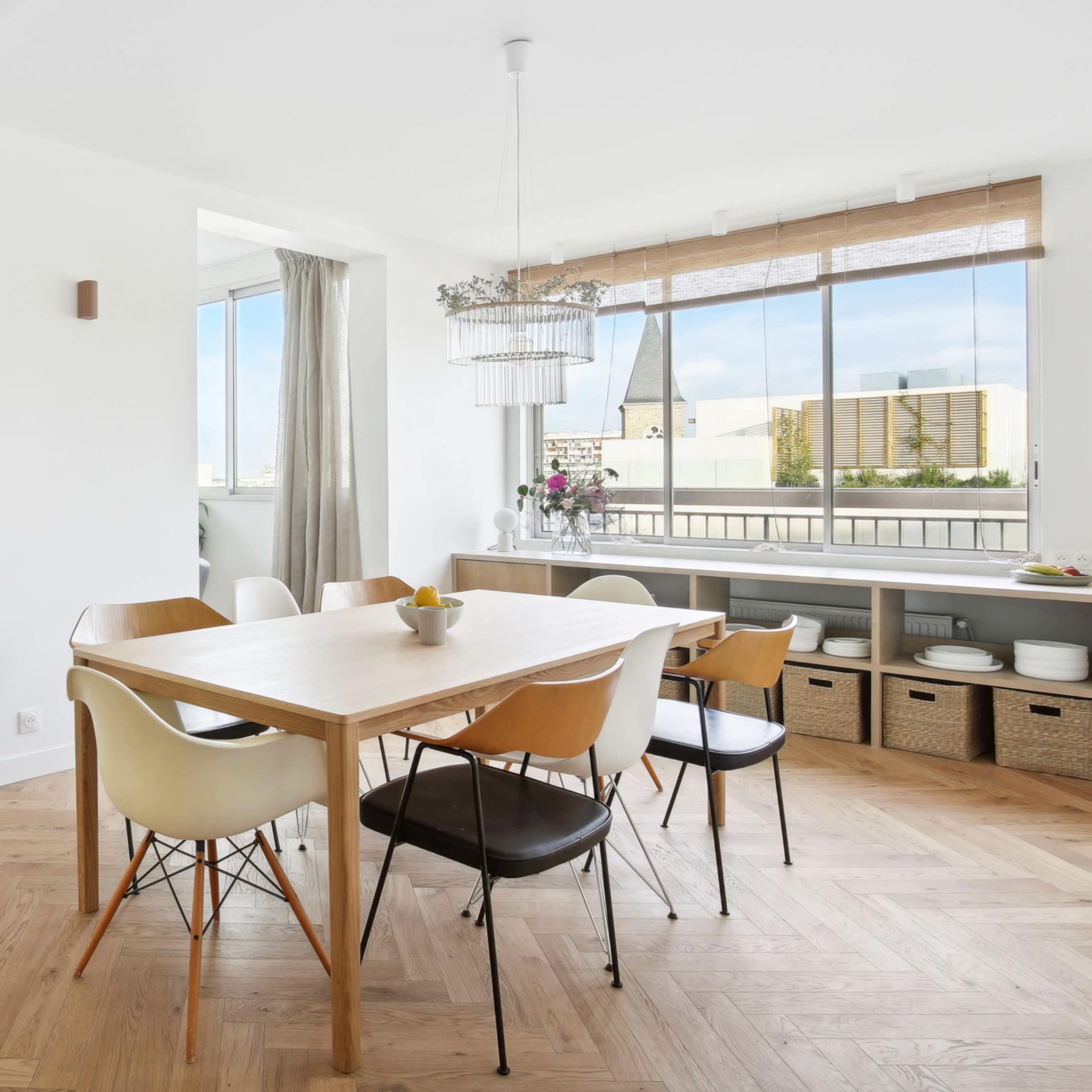 A light-filled dining area features a wooden table surrounded by a mix of chairs, with large windows providing a view of the outdoors.