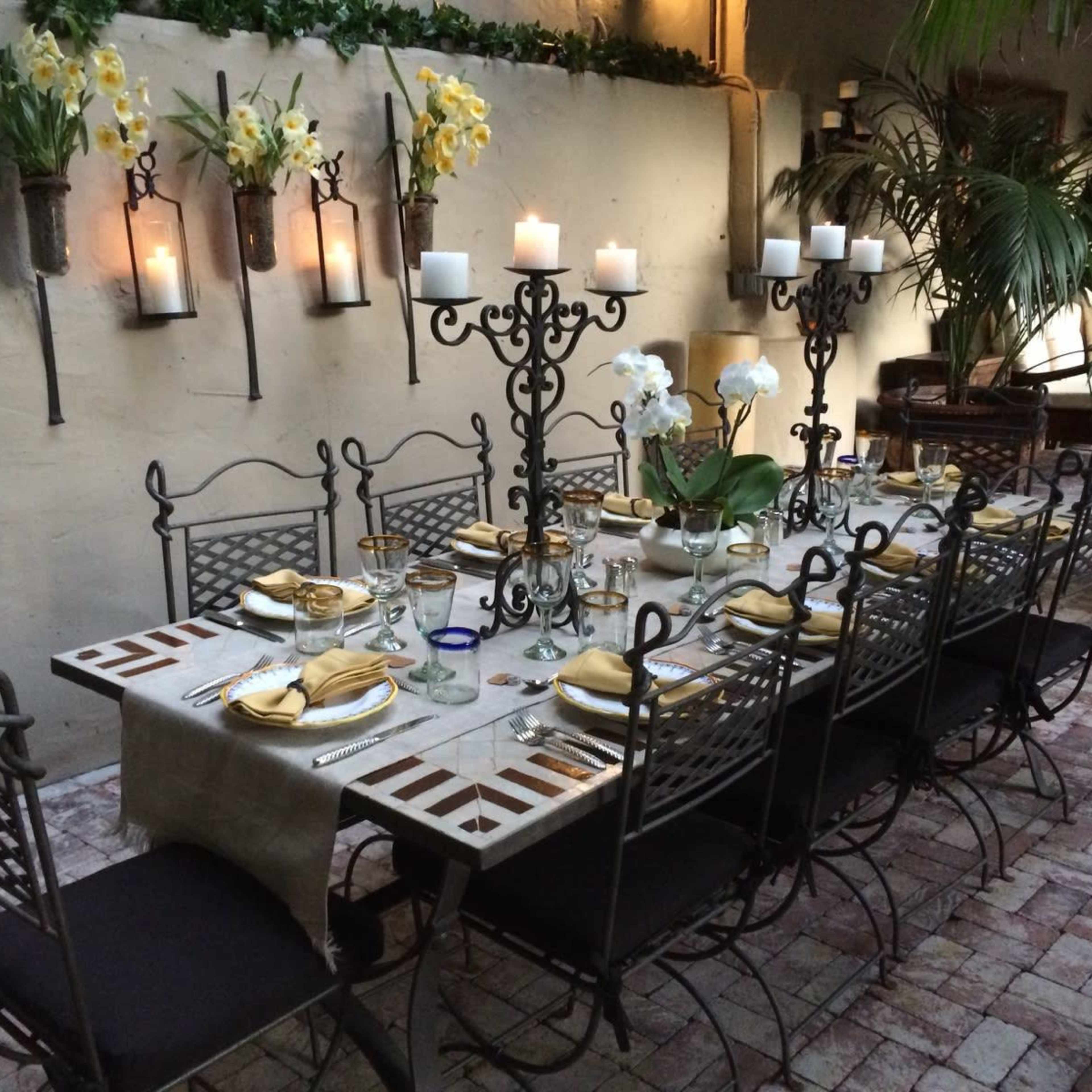 A long, decorated dining table with elegant place settings and tall candle holders is set in a courtyard surrounded by plants and walls adorned with flowers.