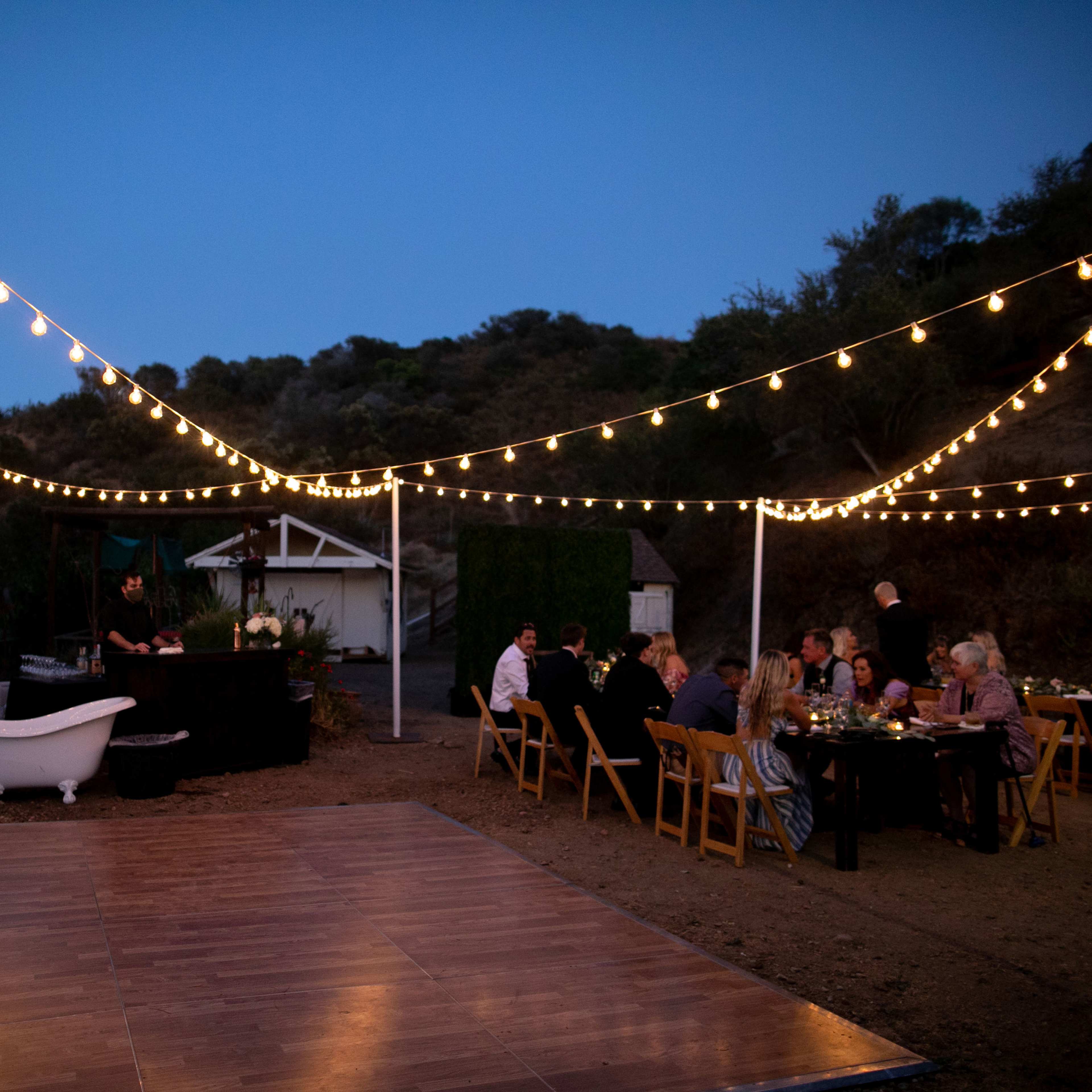 A dinner party is set up outdoors with string lights illuminating the area, featuring a long table and a black serving station, against a backdrop of hills and trees.