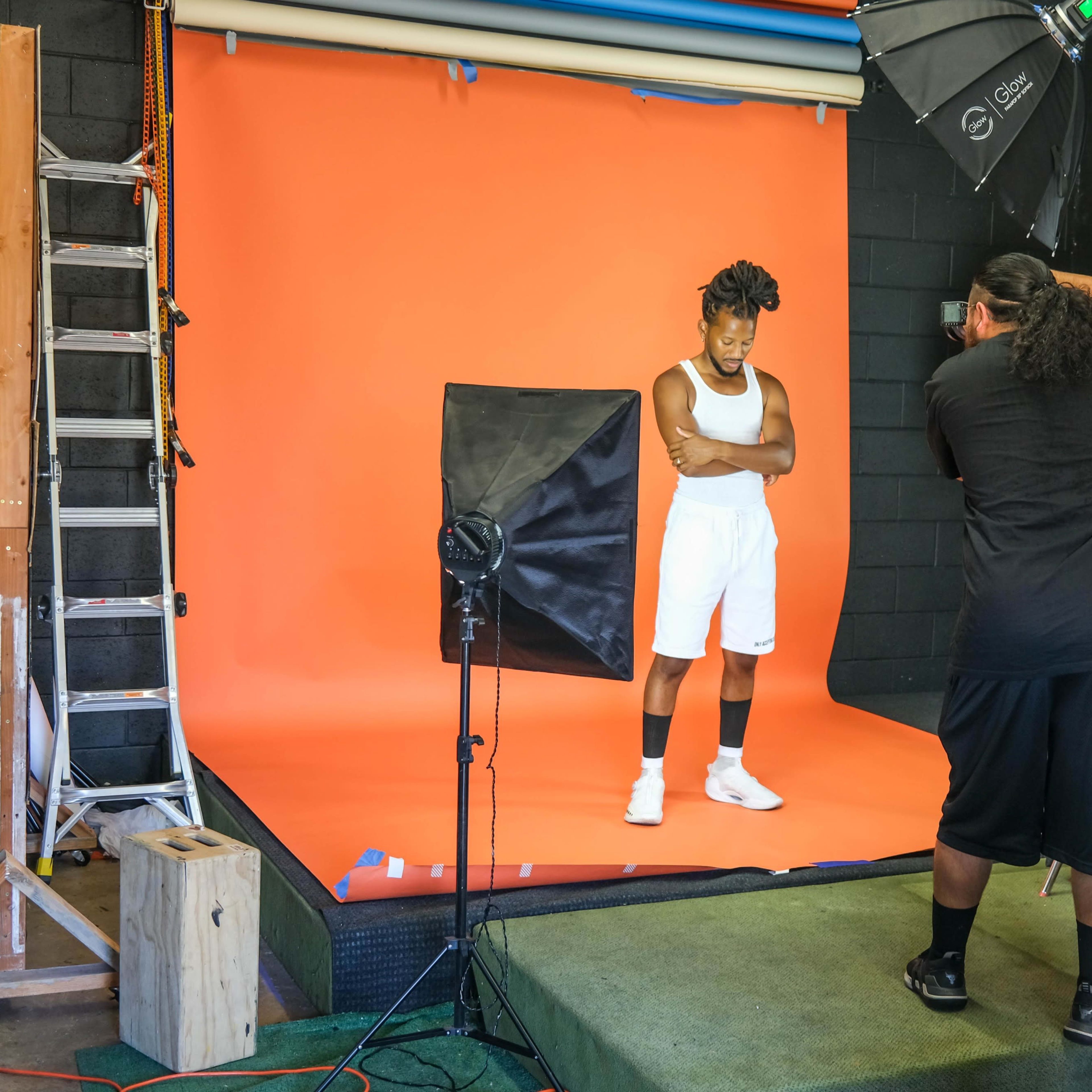 A person poses in a studio setting with an orange backdrop while a photographer adjusts the lighting and equipment.