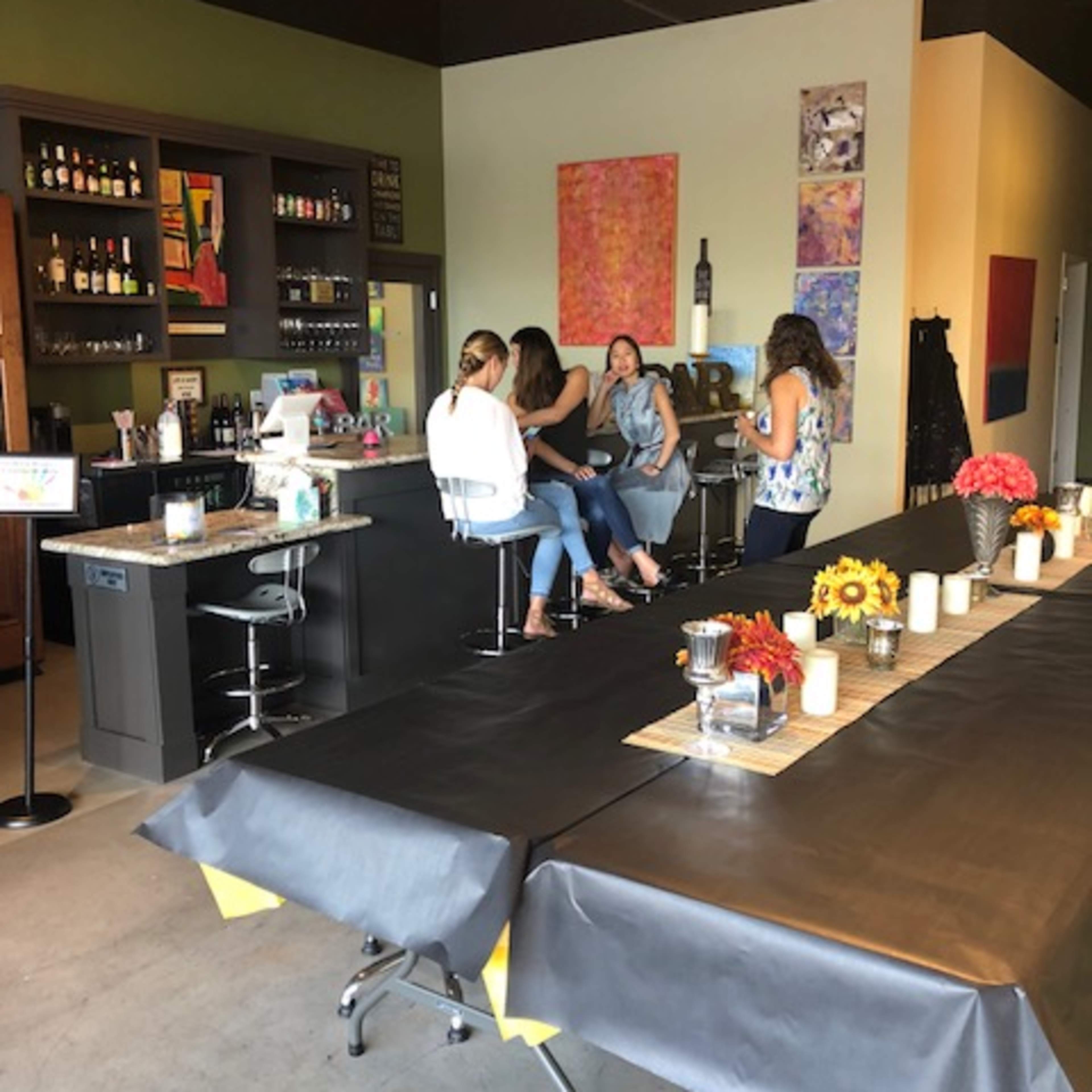 A modern bar area with a long table set for an event and several women seated at the counter engaging in conversation.