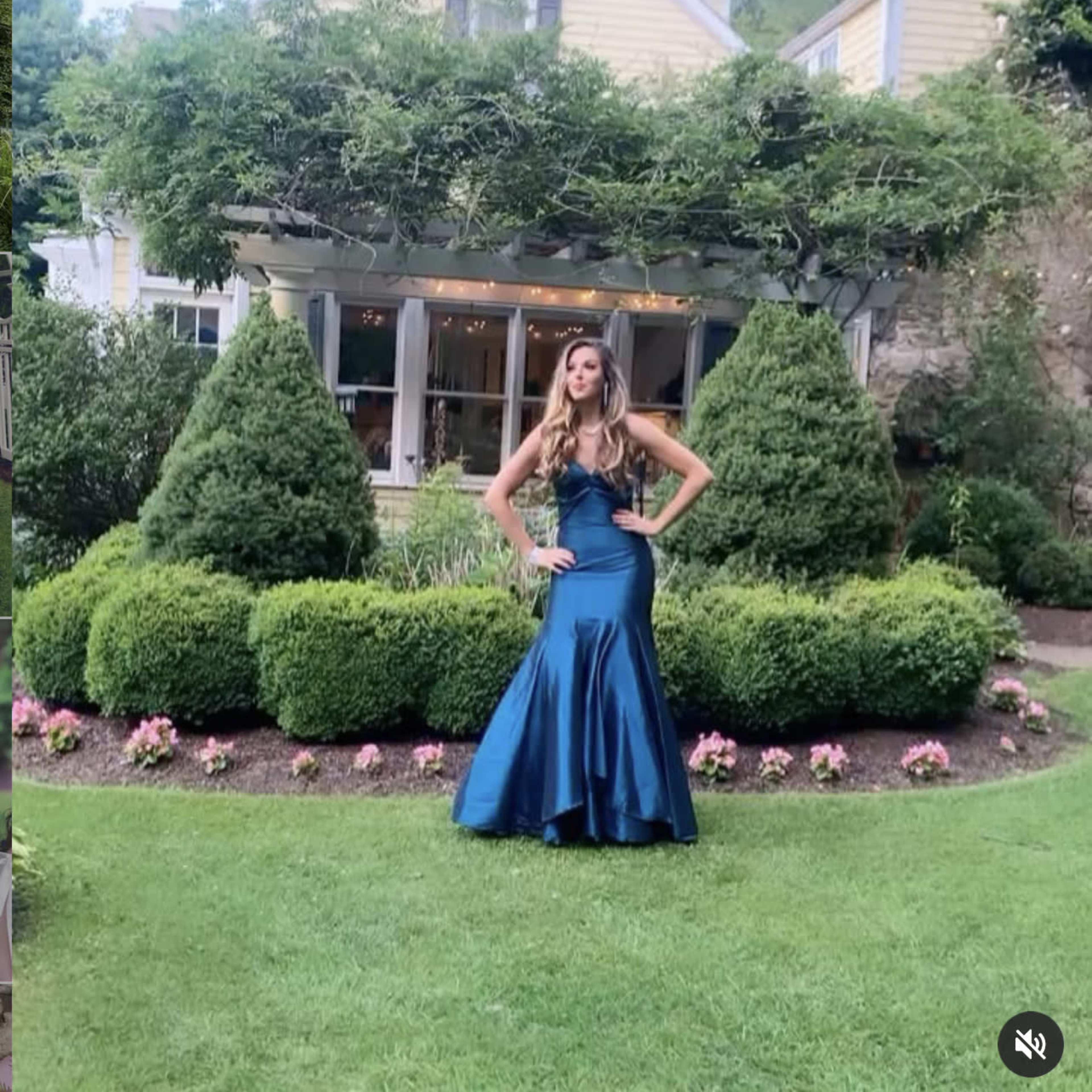 A woman in a blue gown poses in front of a house with landscaped gardens and topiary bushes.