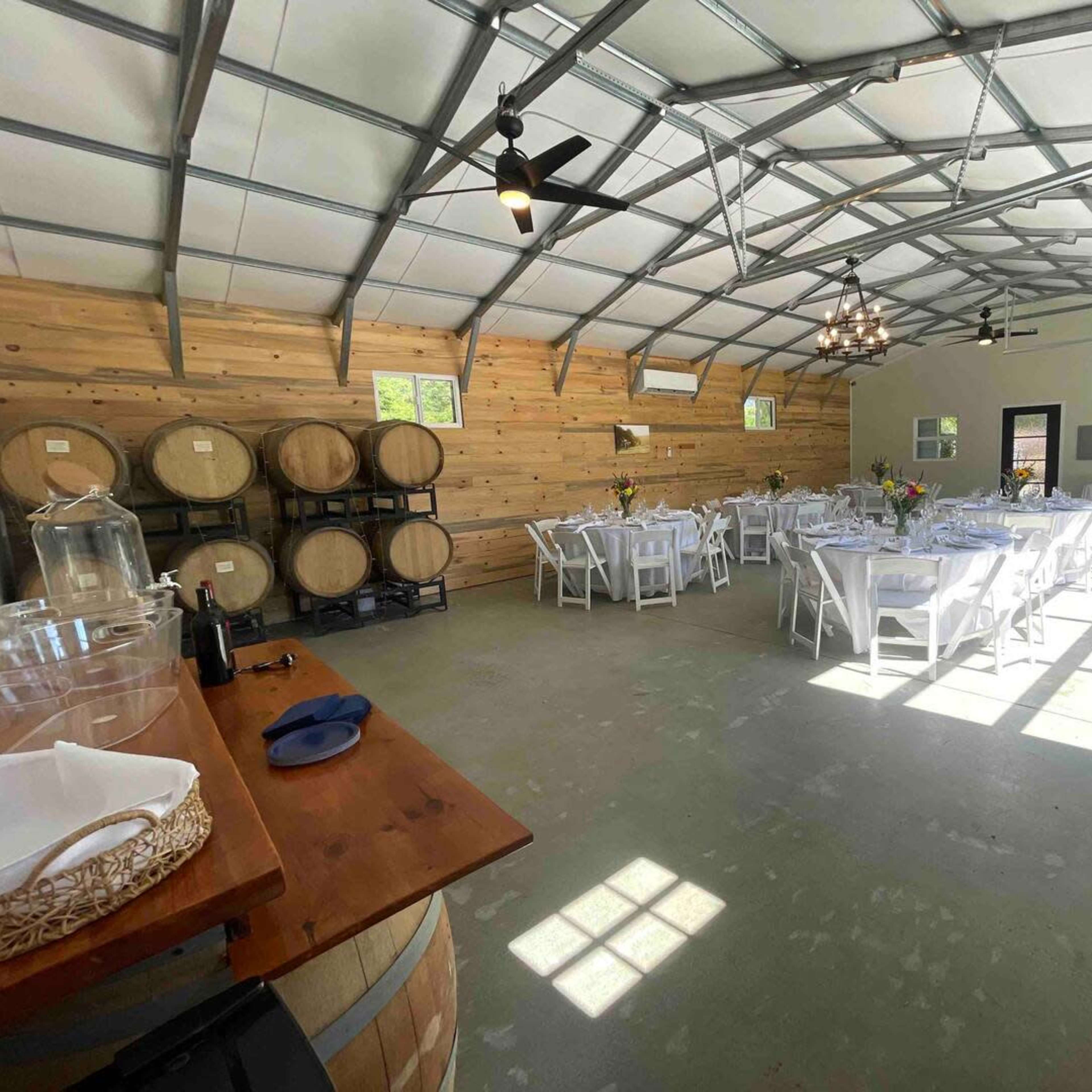 An indoor event space with white tables set for dining, wooden barrels stacked against a wall, and large windows allowing natural light to enter.