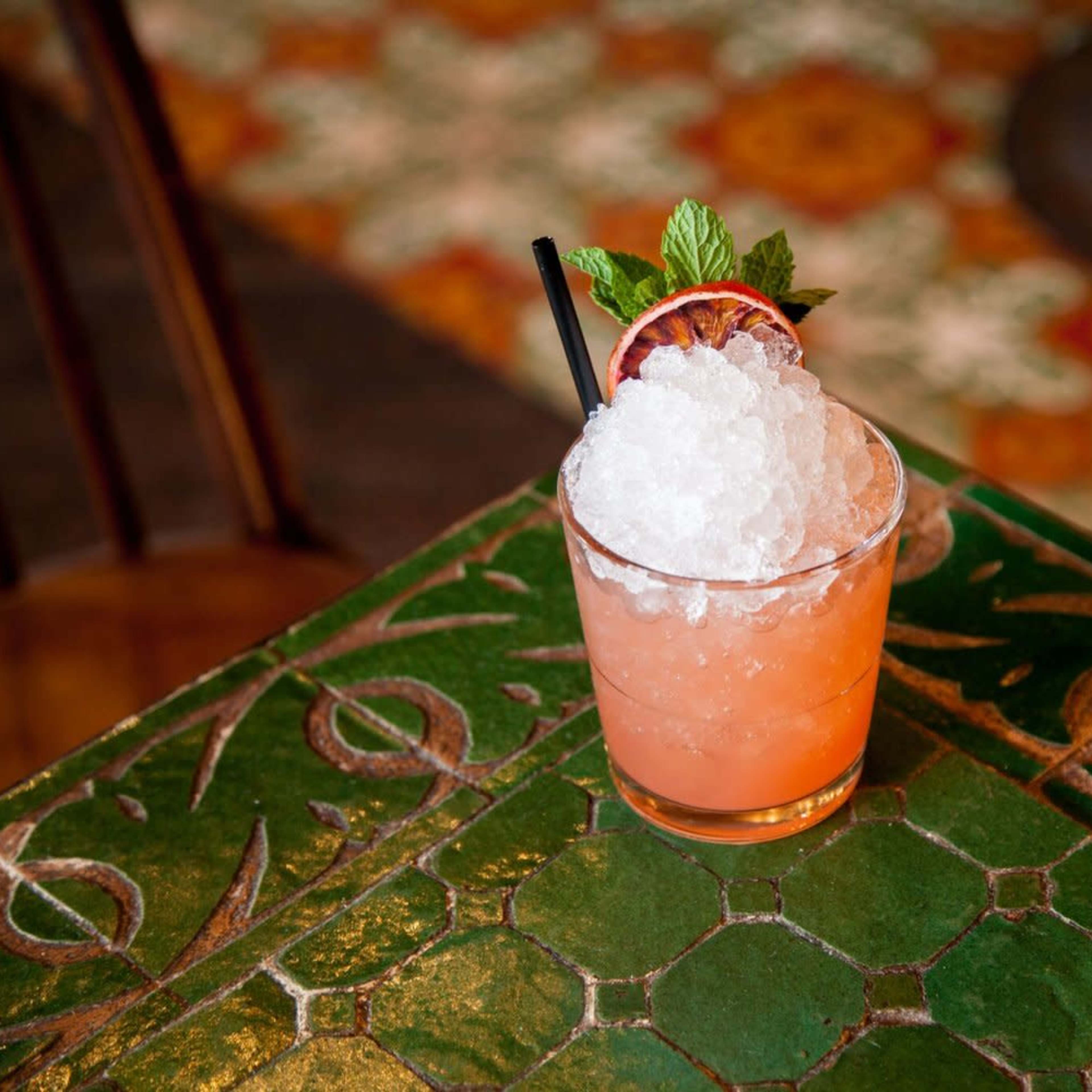 A cocktail filled with crushed ice and garnished with a slice of blood orange and a sprig of mint sits on a green patterned table.