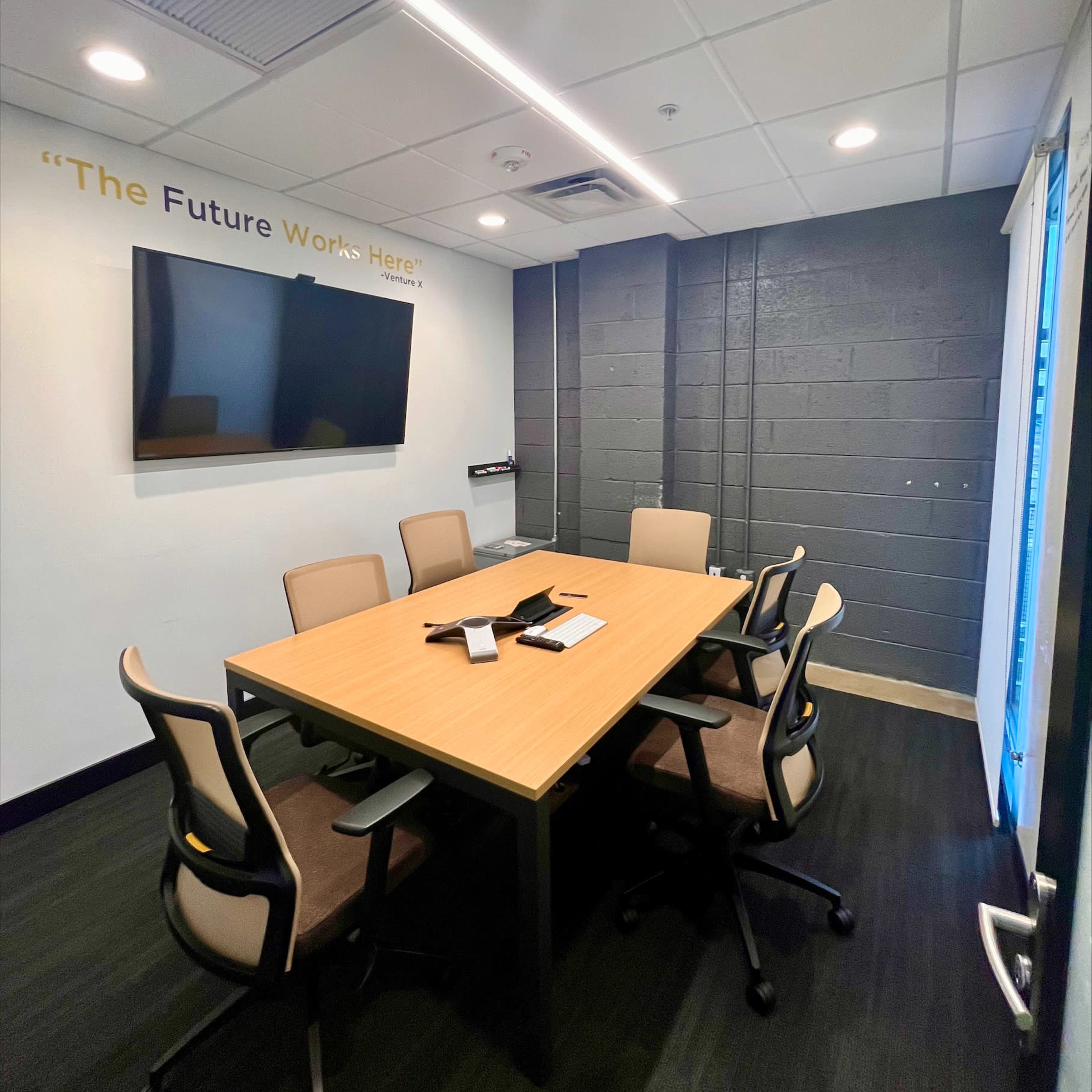 A modern conference room features a large wooden table surrounded by six chairs, a wall-mounted screen, and a slogan on the wall.