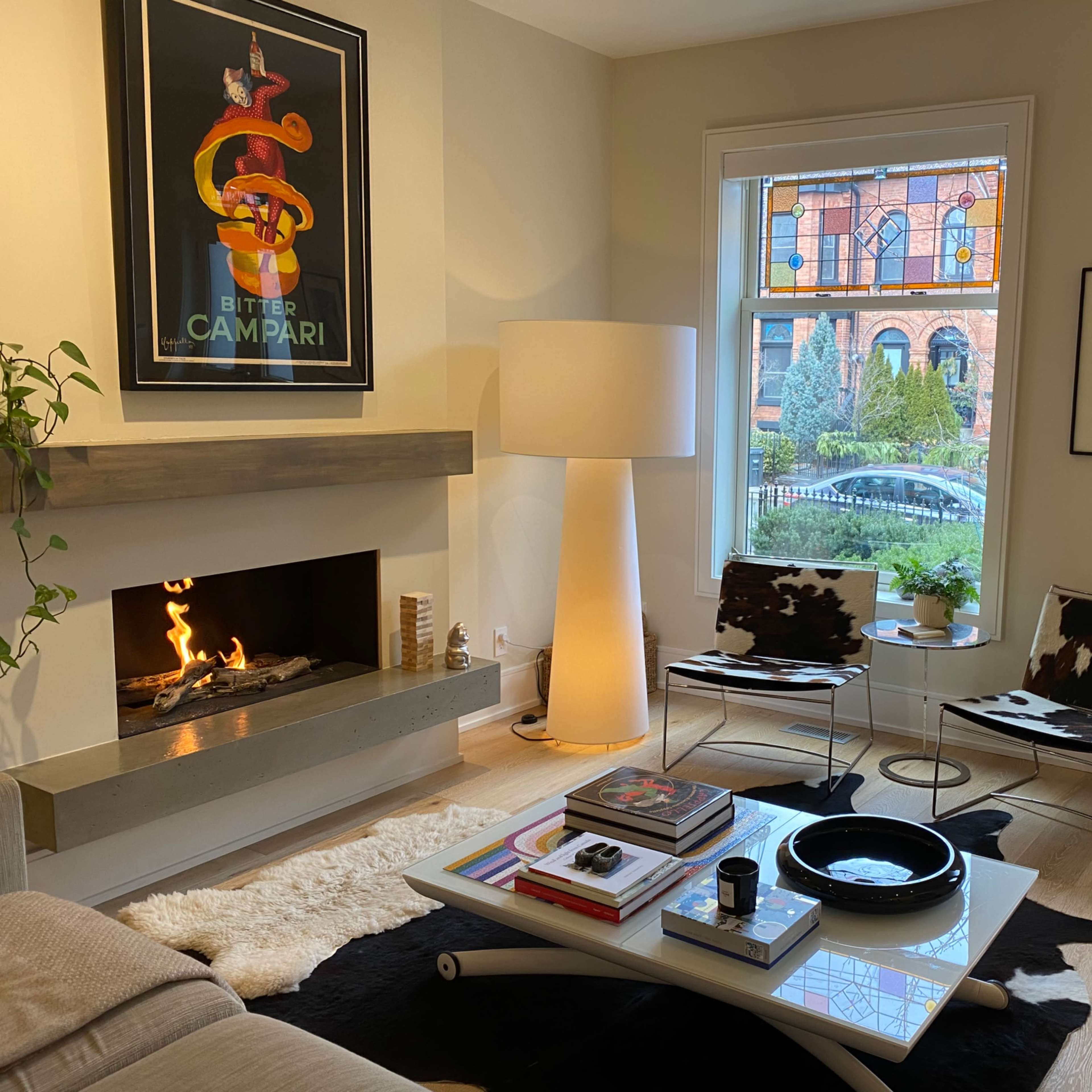 The living room features a modern fireplace, cowhide chairs, a coffee table with books, and a large lamp, with a colorful stained glass window visible outside.