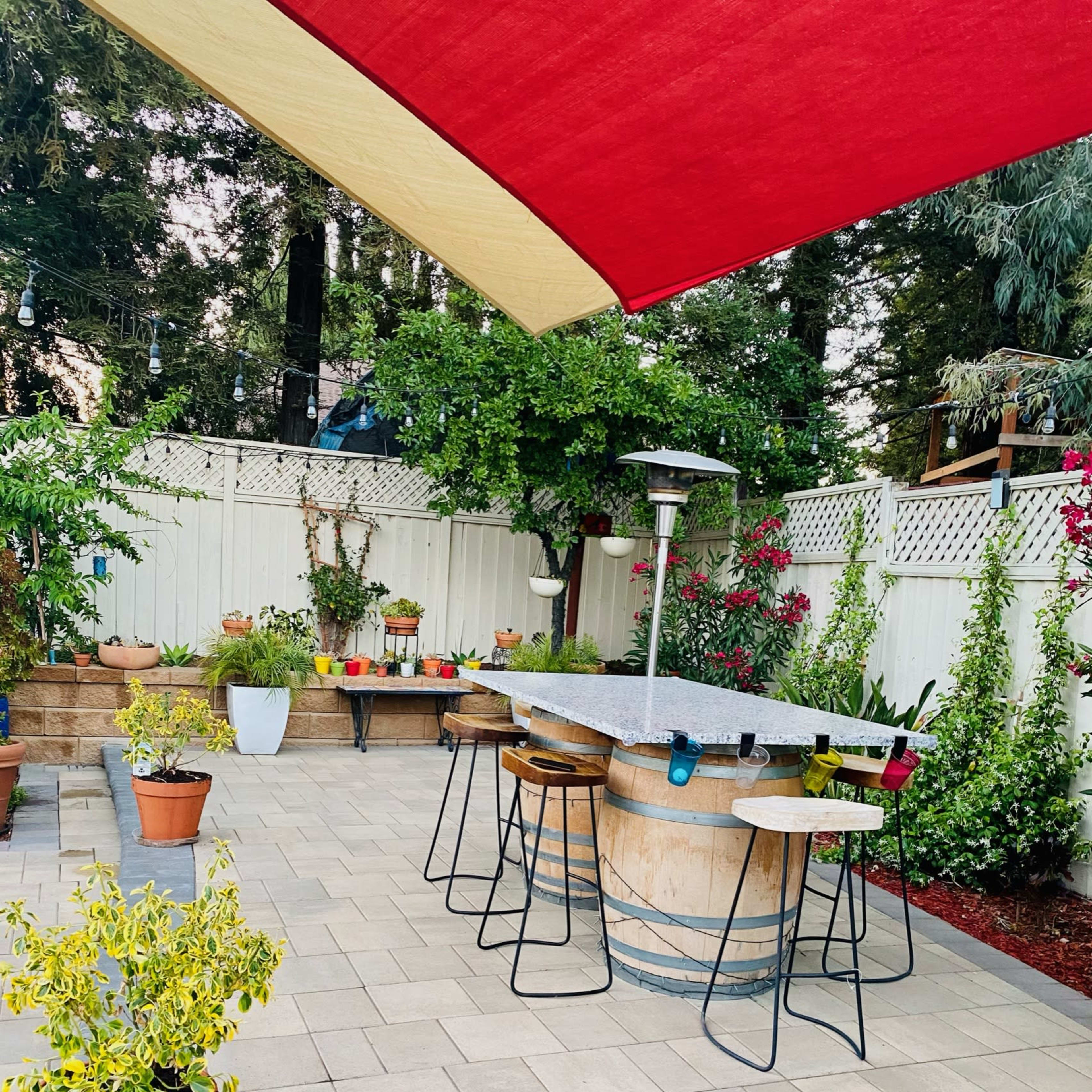 A patio area features a large round table surrounded by tall stools, with several potted plants and a shaded canopy overhead.
