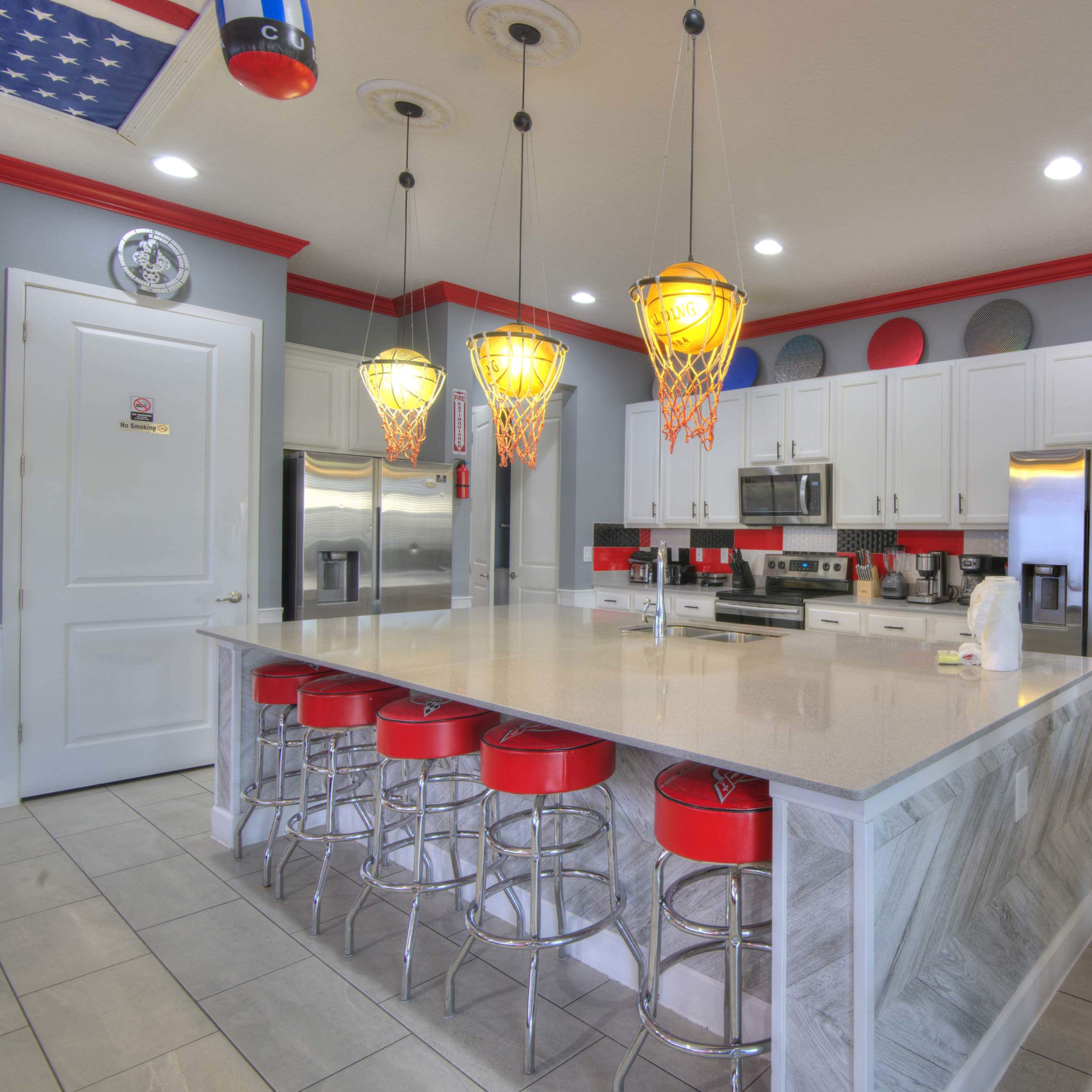 A modern kitchen with a large island, red bar stools, pendant lights shaped like basketball hoops, and stainless steel appliances.