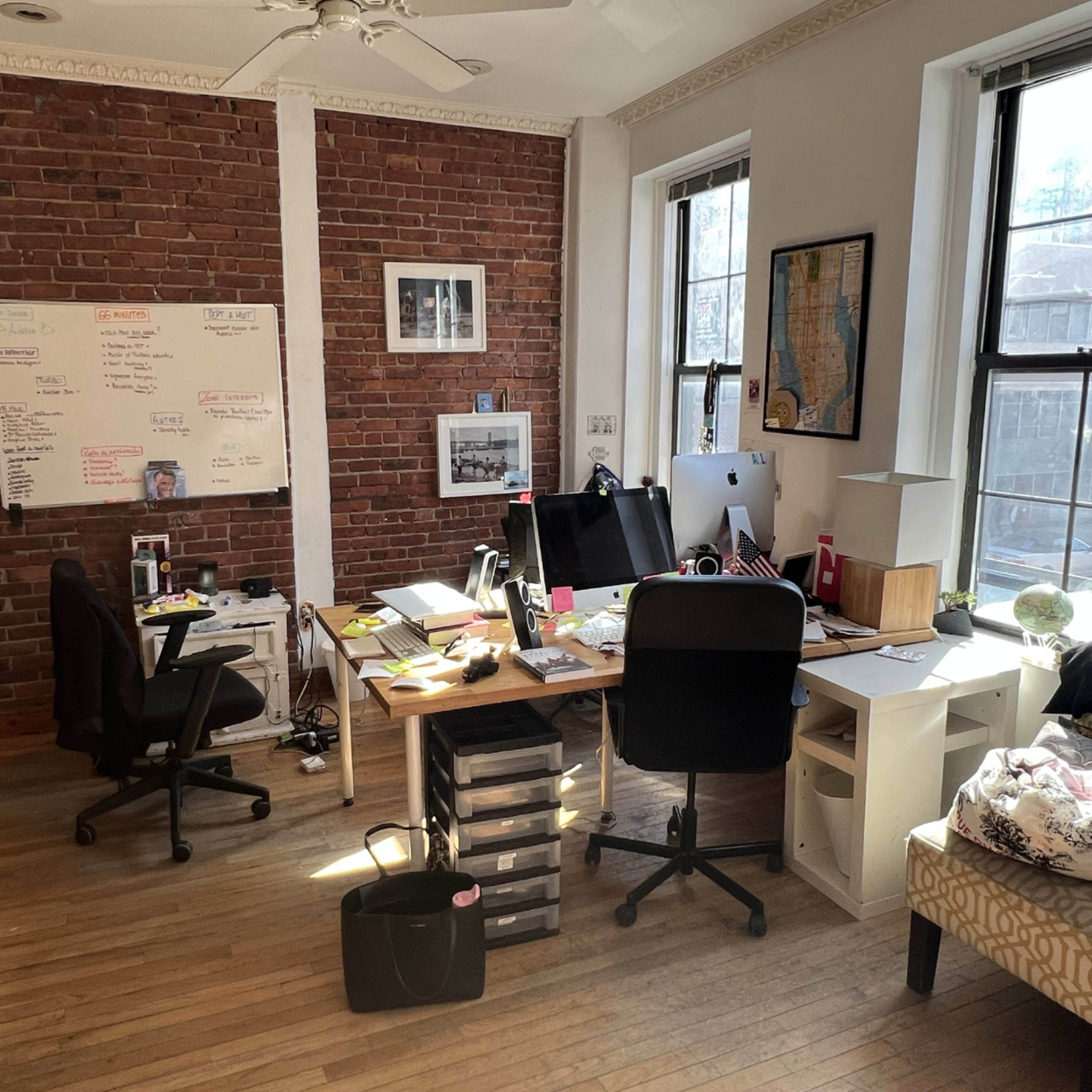 A modern office space with a wooden floor, brick walls, and a desk cluttered with various office supplies and electronic devices.