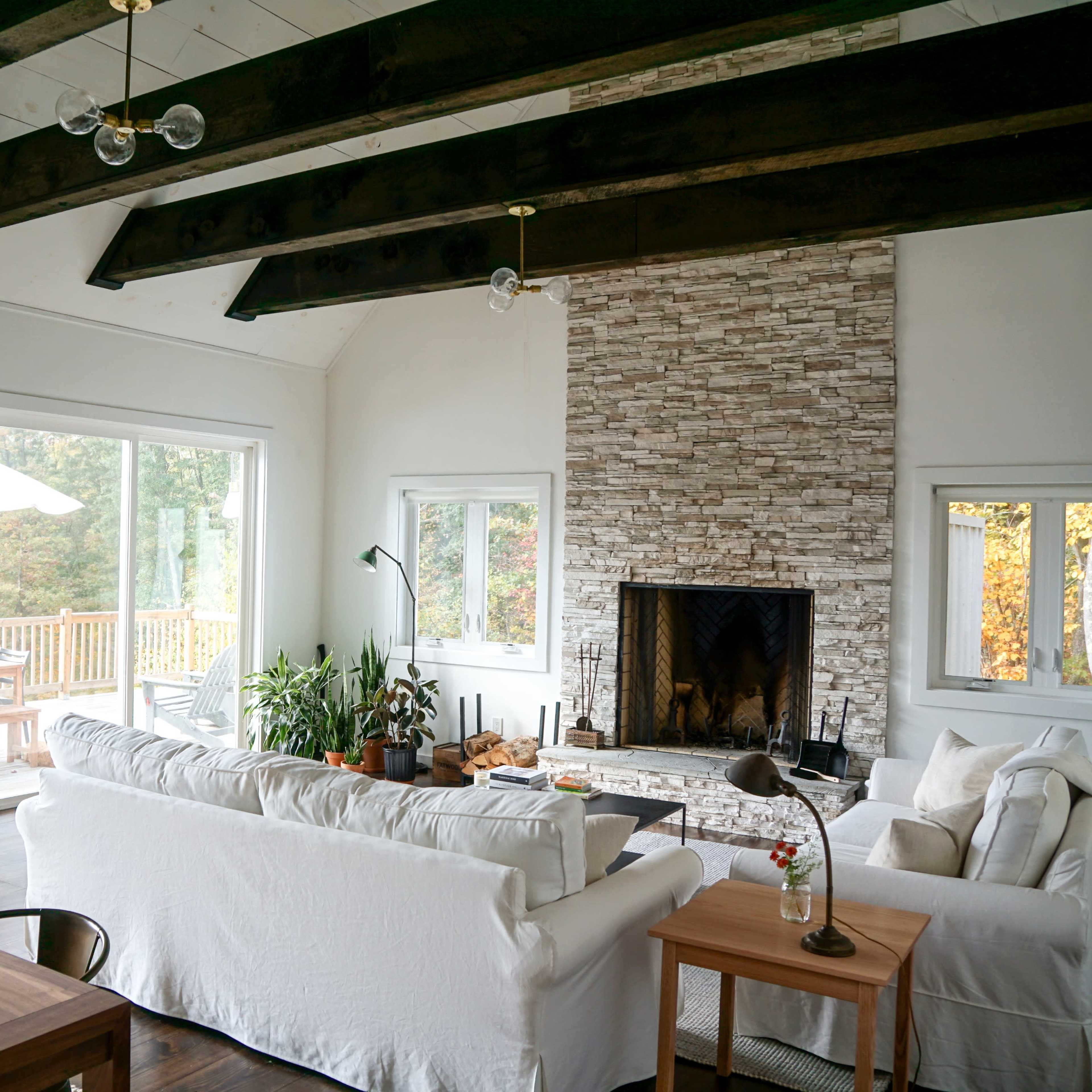 A modern living room with a stone fireplace, large windows, and wooden beams across the ceiling.