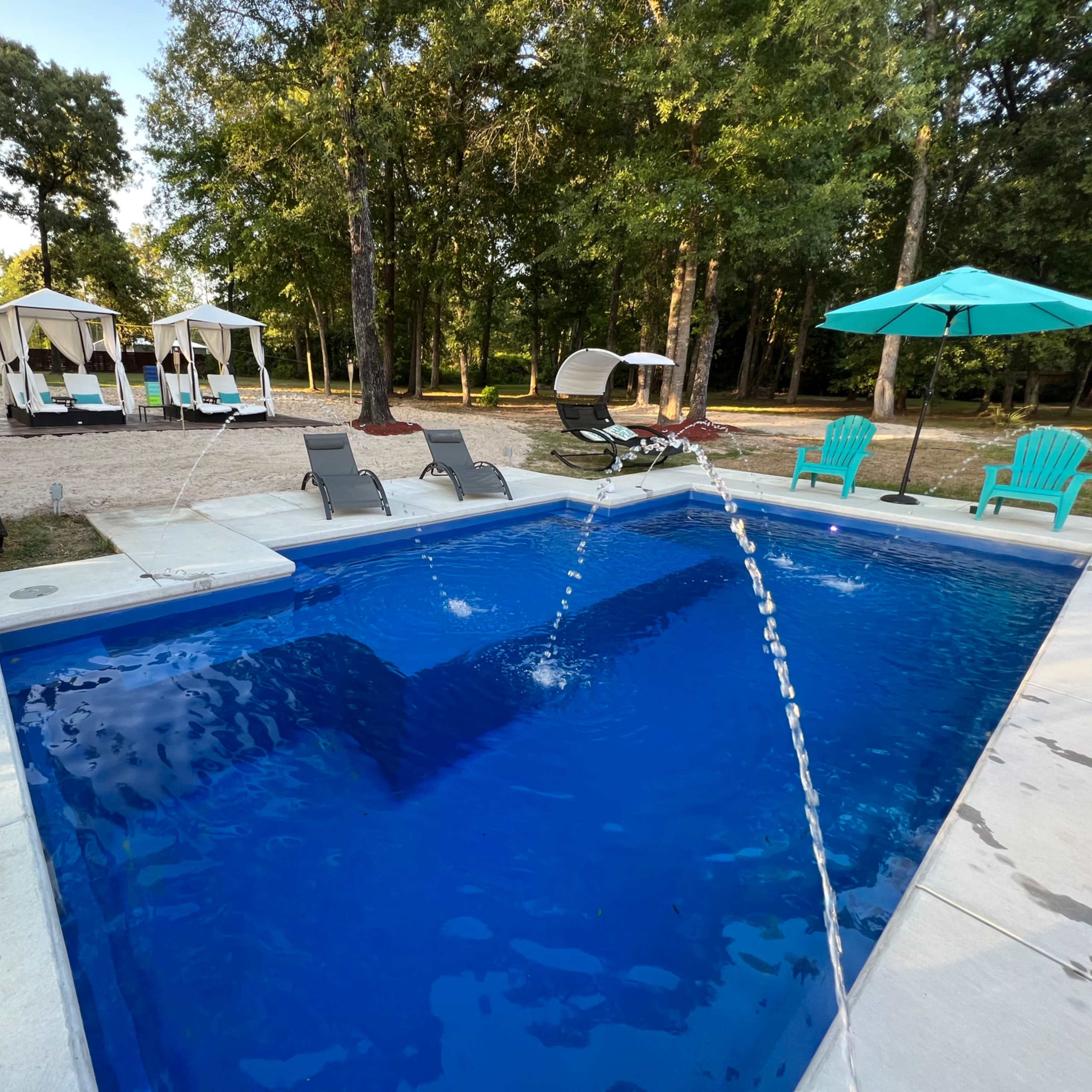 A swimming pool surrounded by lounge chairs and shaded cabanas in a wooded outdoor area.