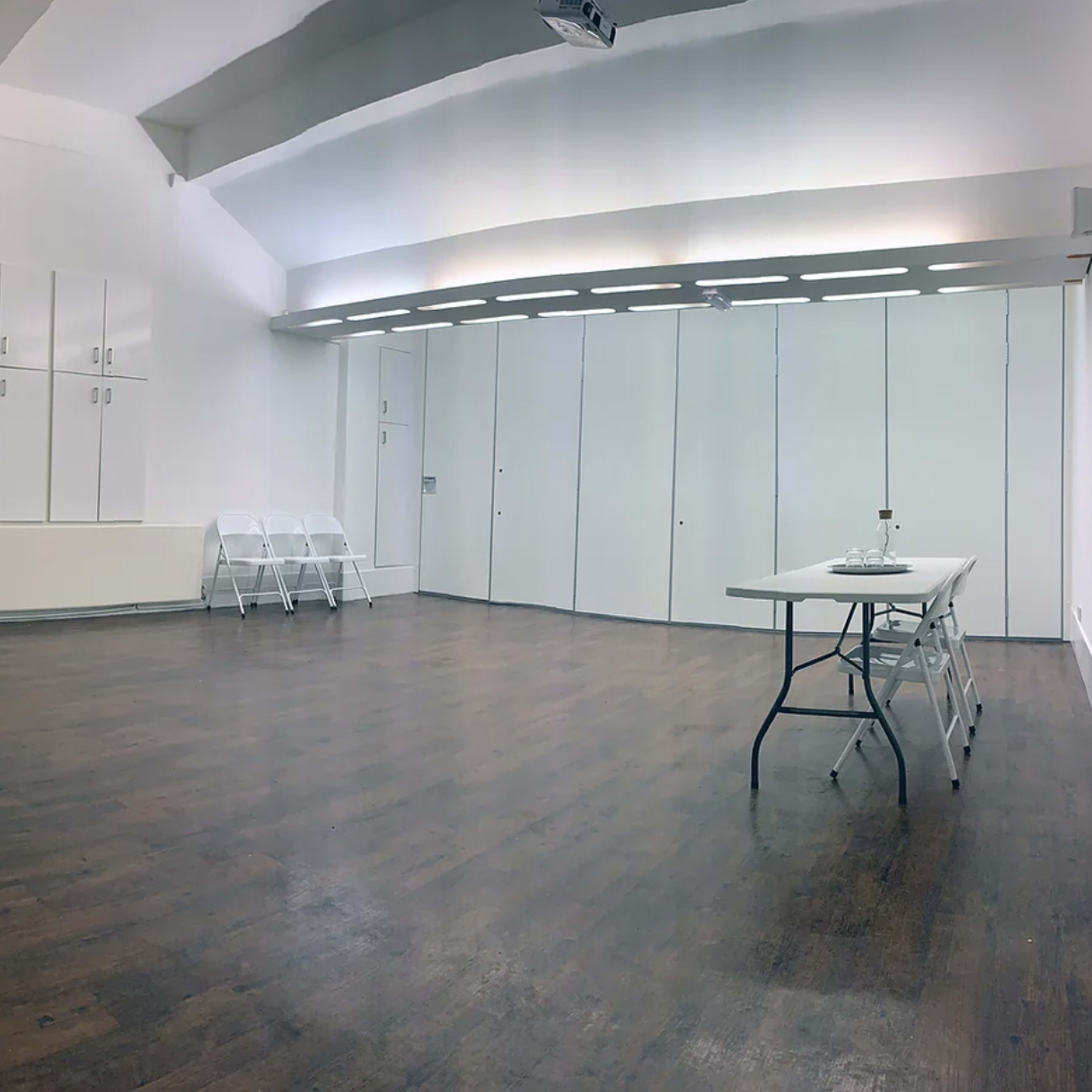 A spacious, empty room with hardwood flooring, white walls, and several folding chairs arranged against one side.
