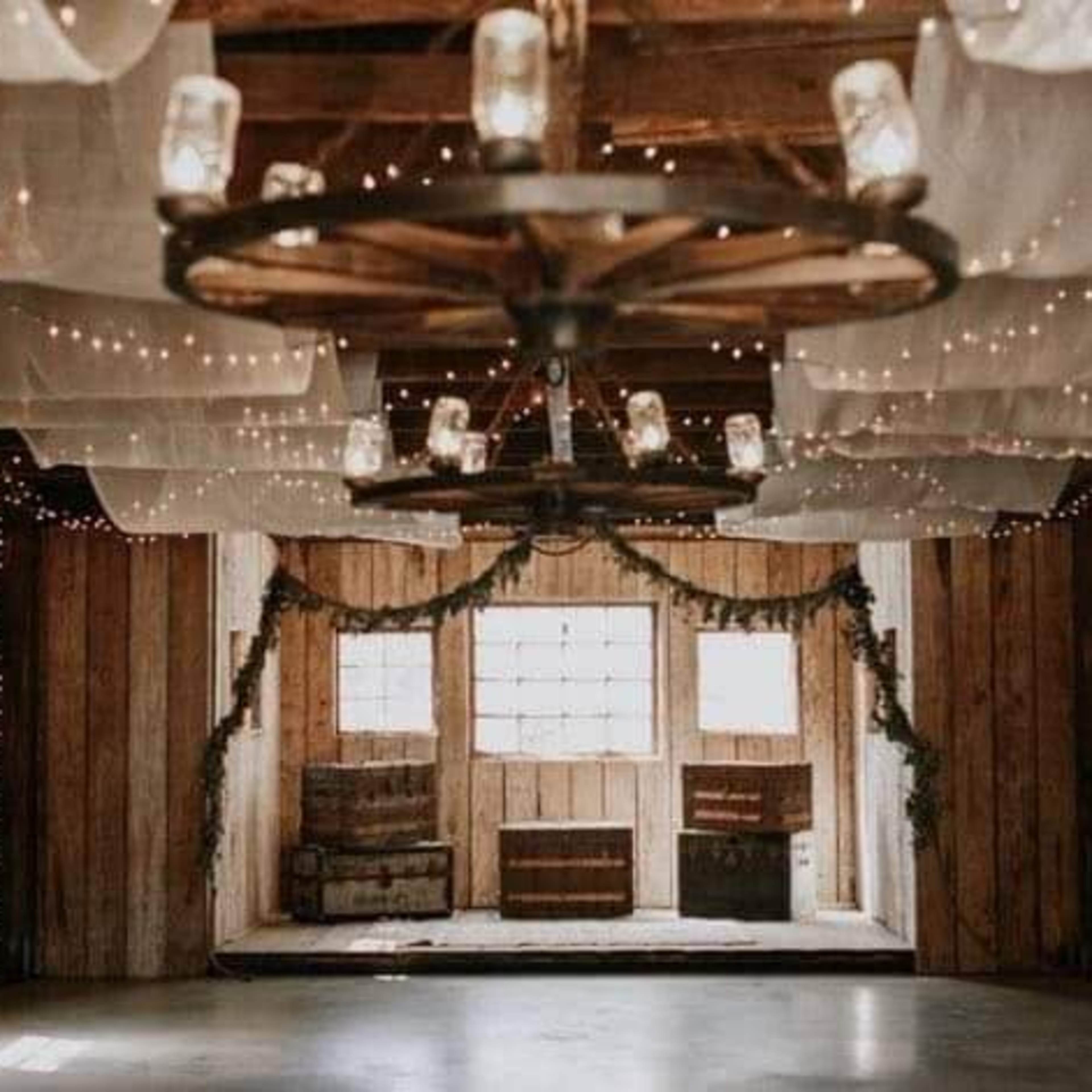 A rustic interior of a barn with wooden walls, a ceiling adorned with lights and draped fabric, and wooden crates positioned in front of large windows.