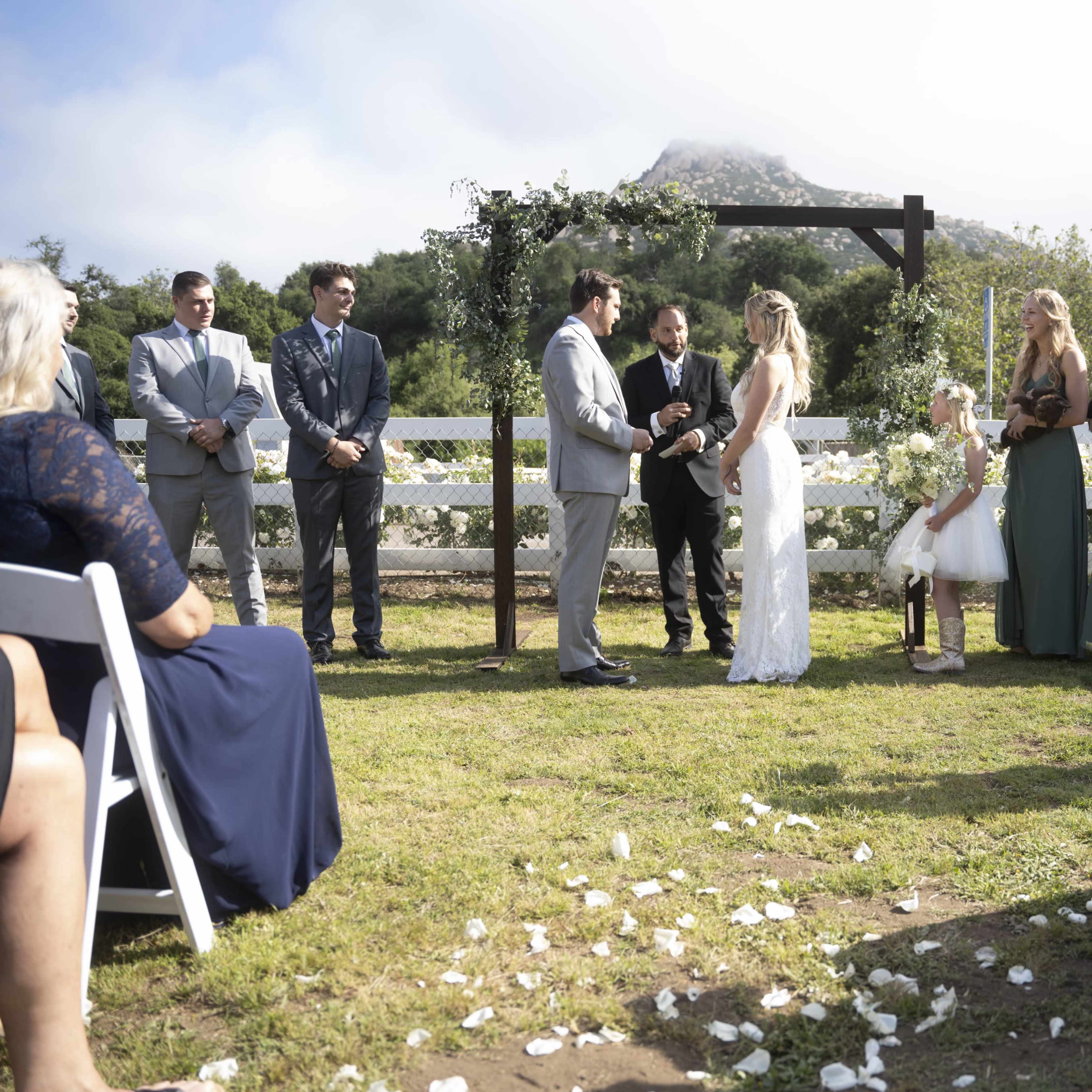 A couple exchanges vows during an outdoor wedding ceremony in front of guests seated on white chairs.