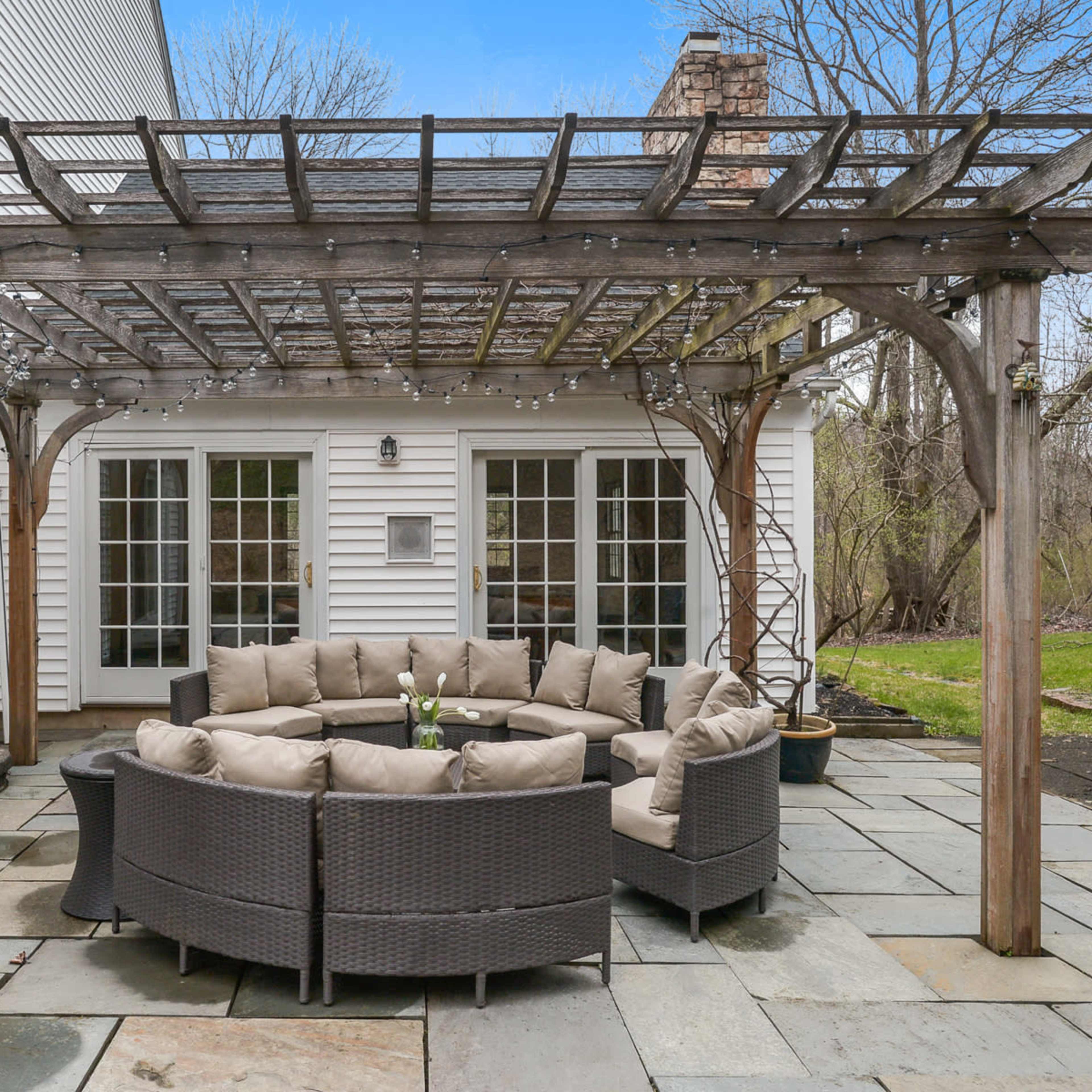 A circular seating area with a couch and table is located under a wooden pergola on a stone patio, surrounded by lush greenery.