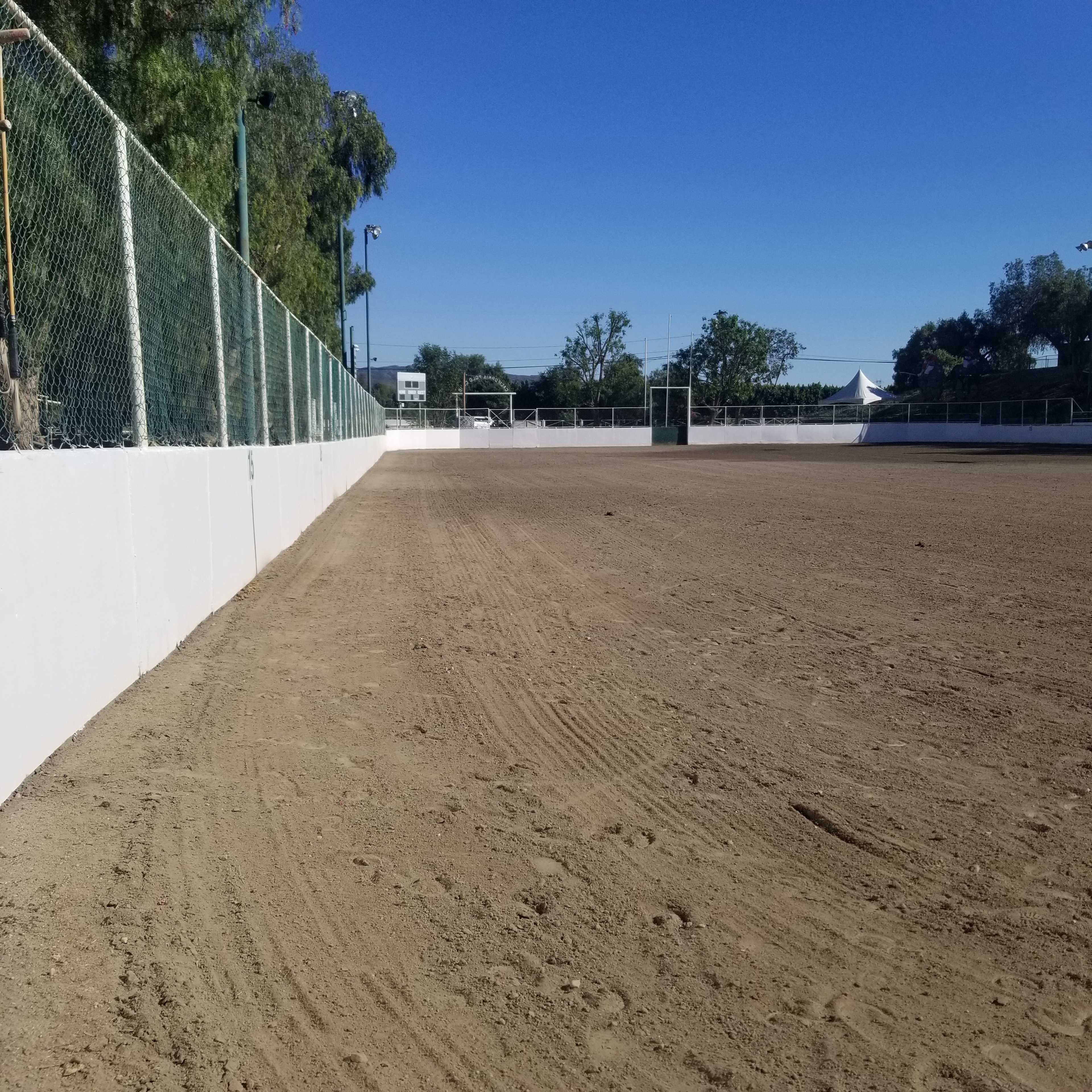 A fenced dirt riding arena with a clear blue sky and a tree line in the background.