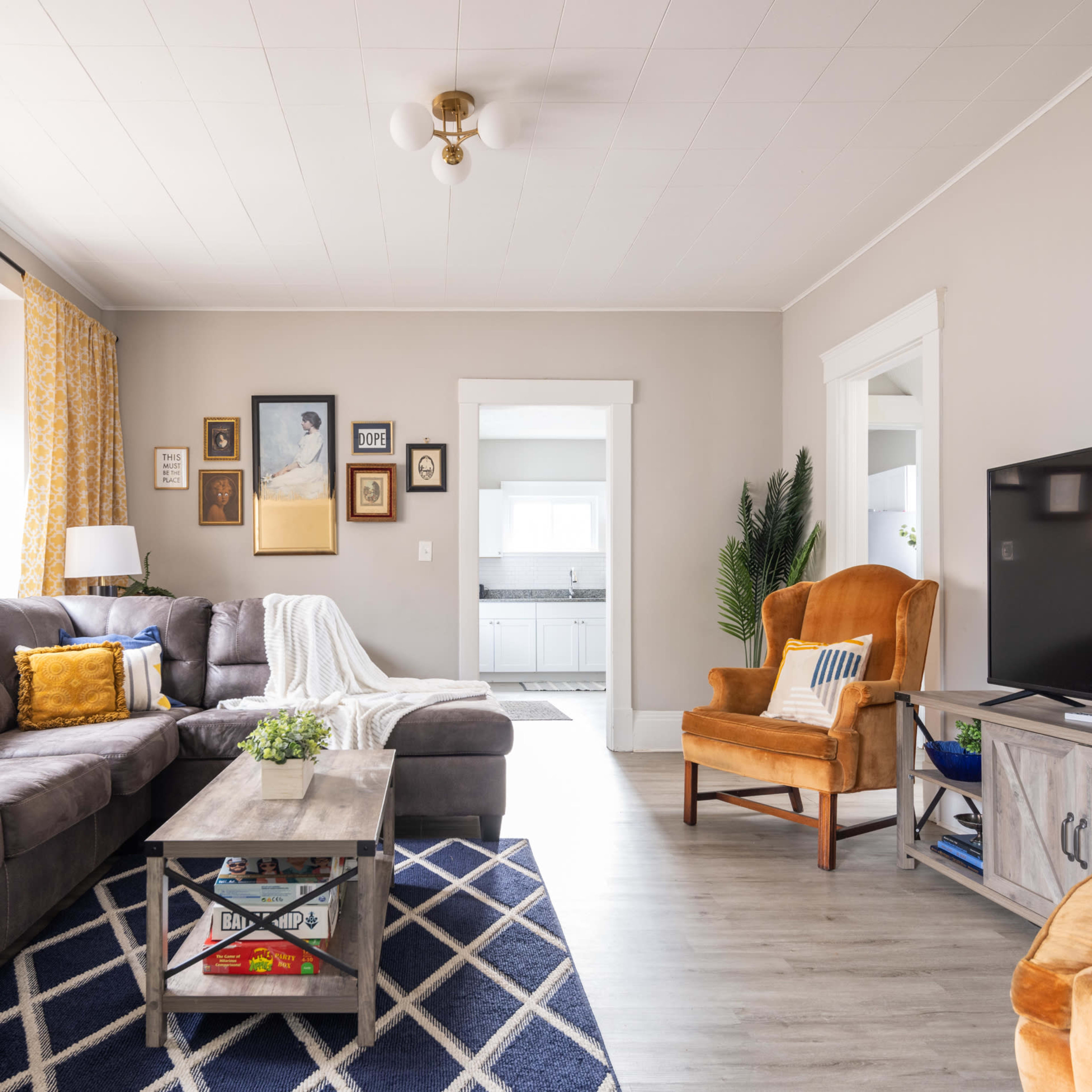 The living room features a gray sectional couch, a wooden coffee table, an orange armchair, and a television against a light-colored wall, with decorative curtains and framed pictures.