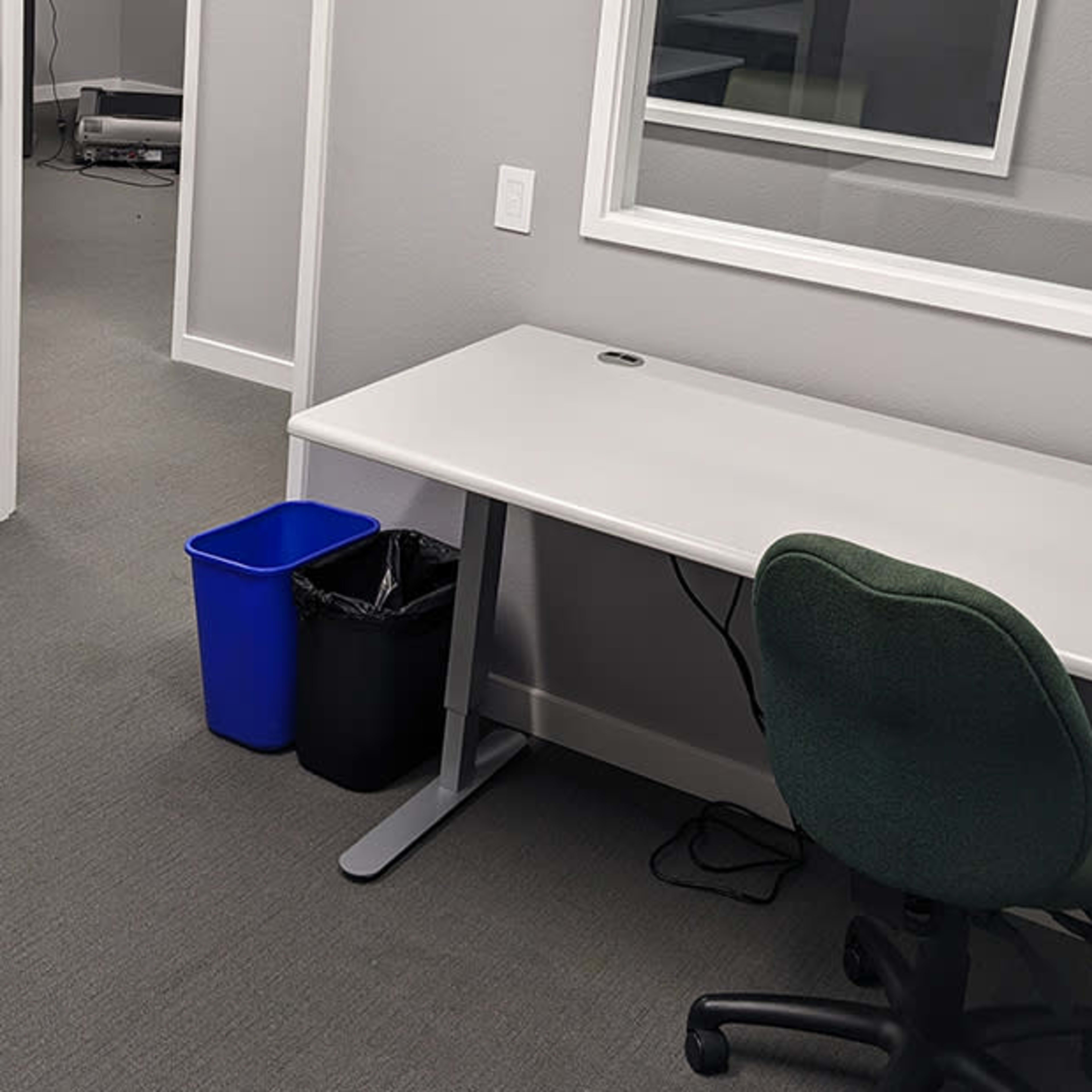 A minimalist office space features two desks, a blue recycling bin, and a green office chair, with a door and a mirrored wall visible.