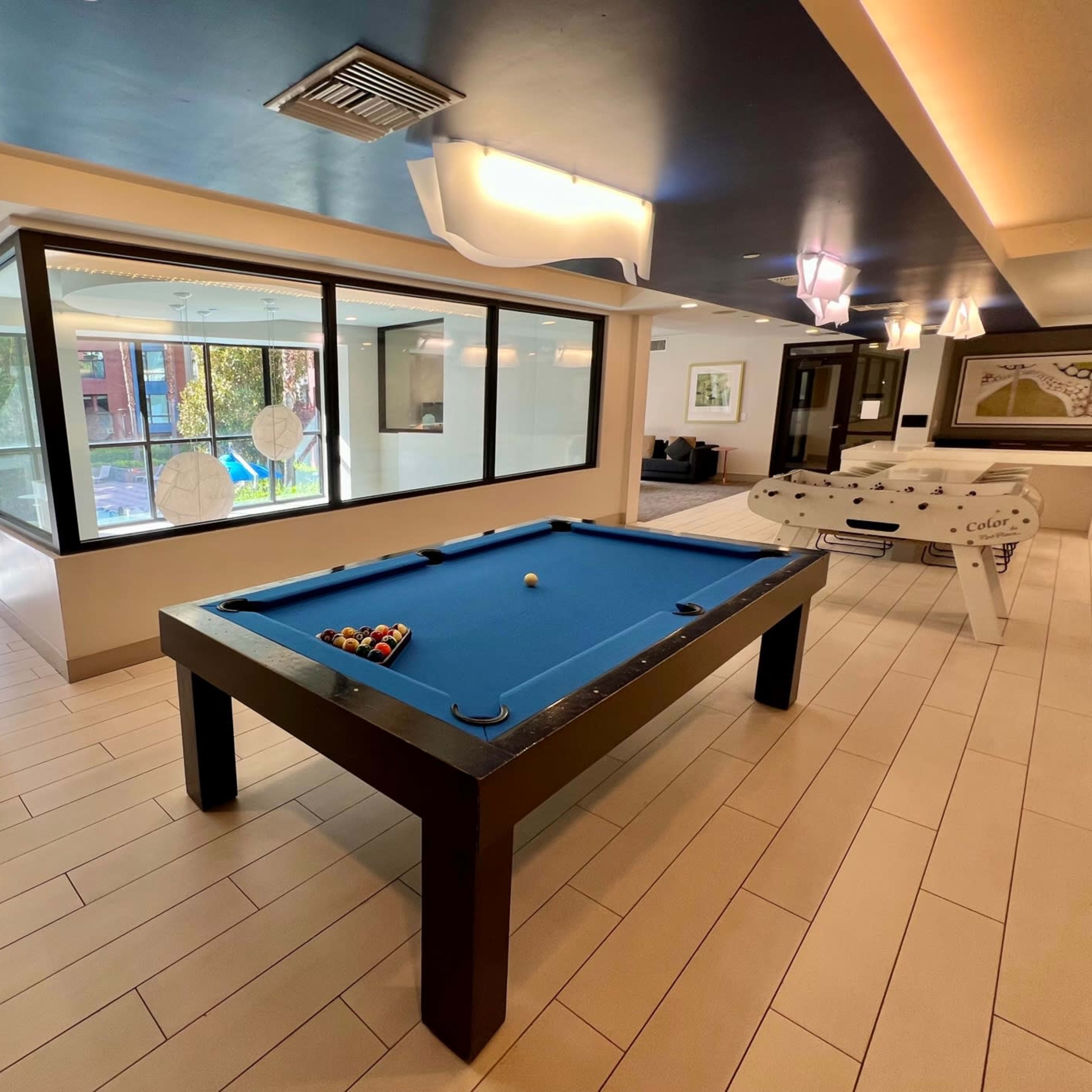 A recreational room featuring a blue billiards table, a foosball table, and large windows with outdoor views.