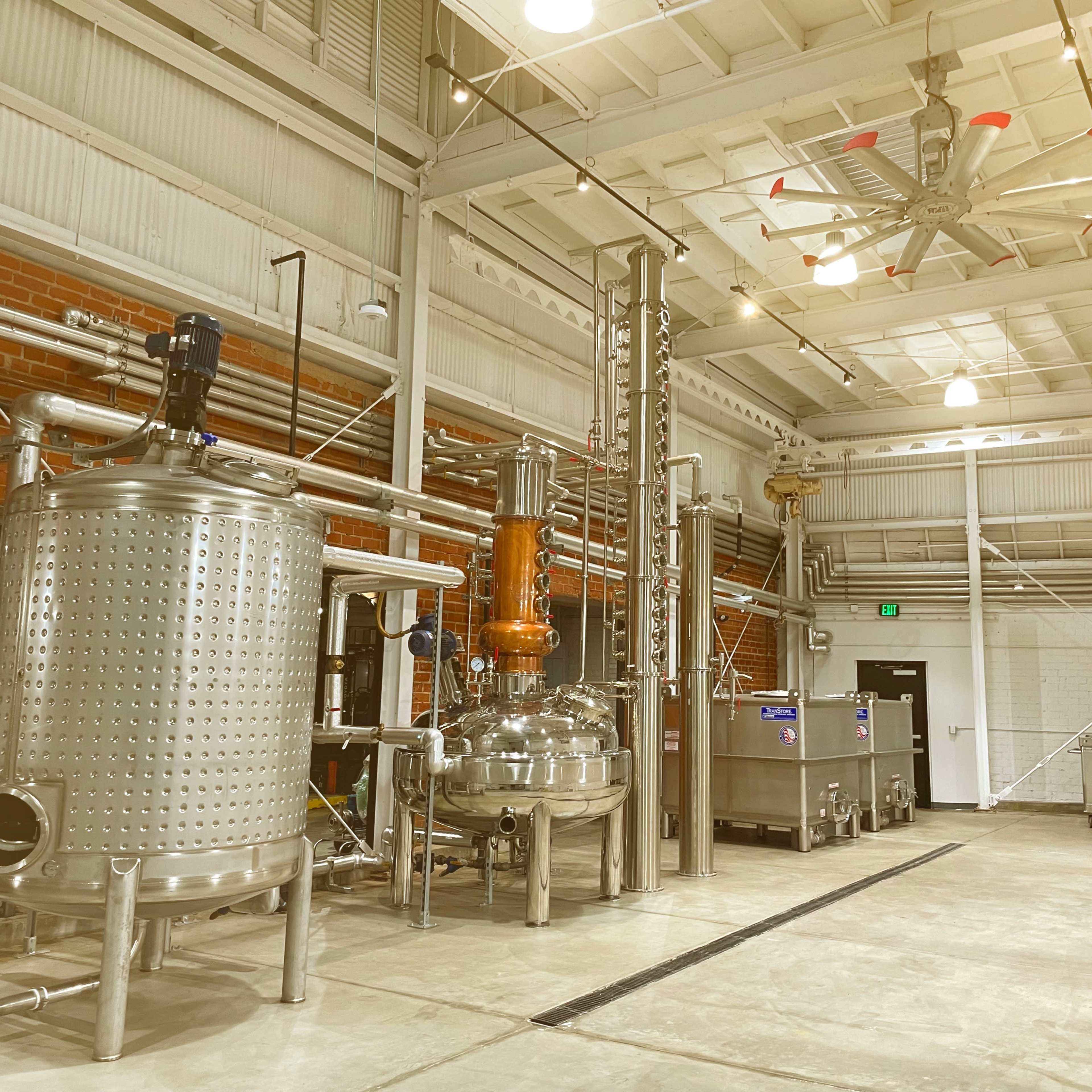 A spacious industrial facility featuring various silver distillation equipment, including a large fermentation tank and a copper still, with exposed brick walls and overhead lighting.