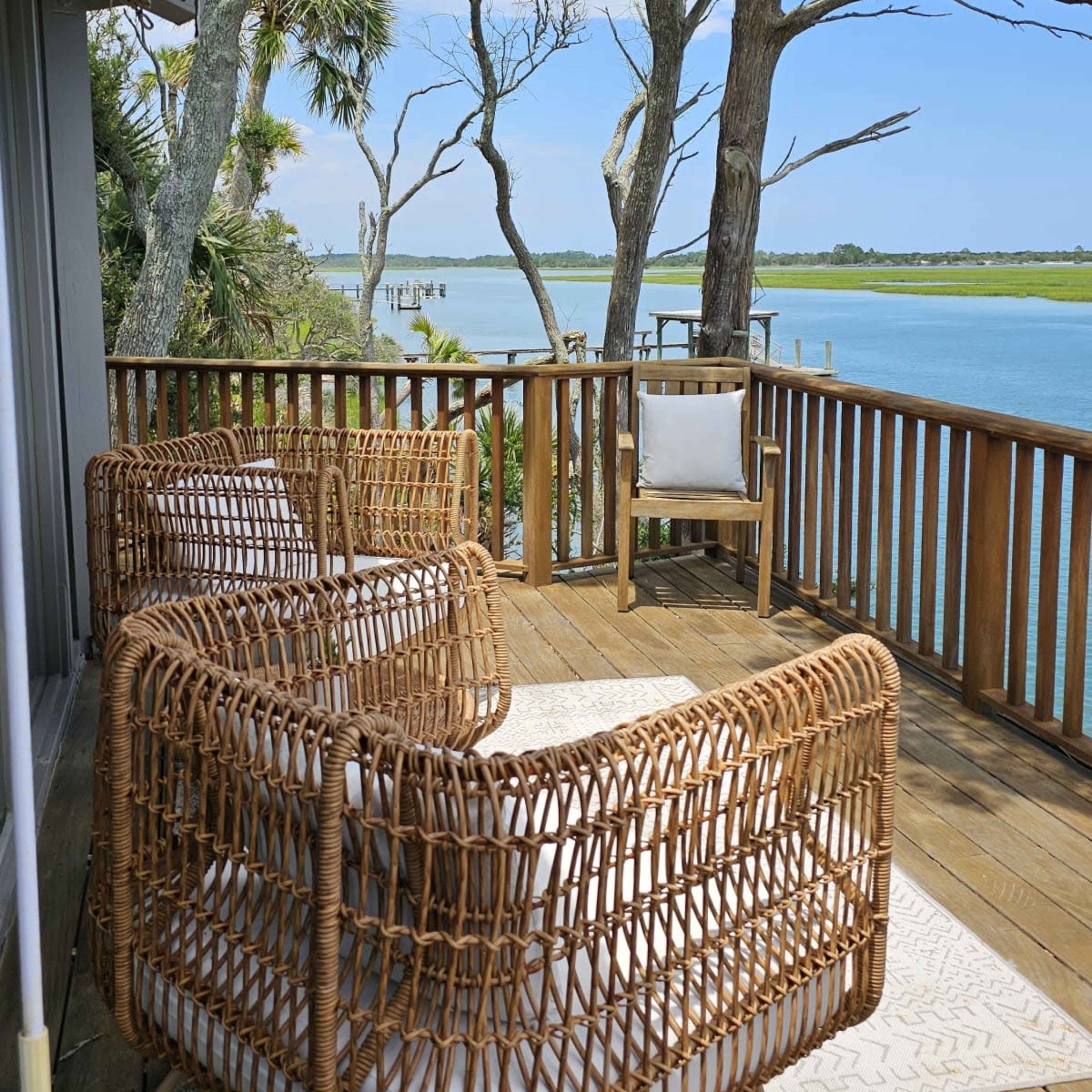 A wooden deck features two rattan chairs and a small table, overlooking a calm body of water and lined by trees.