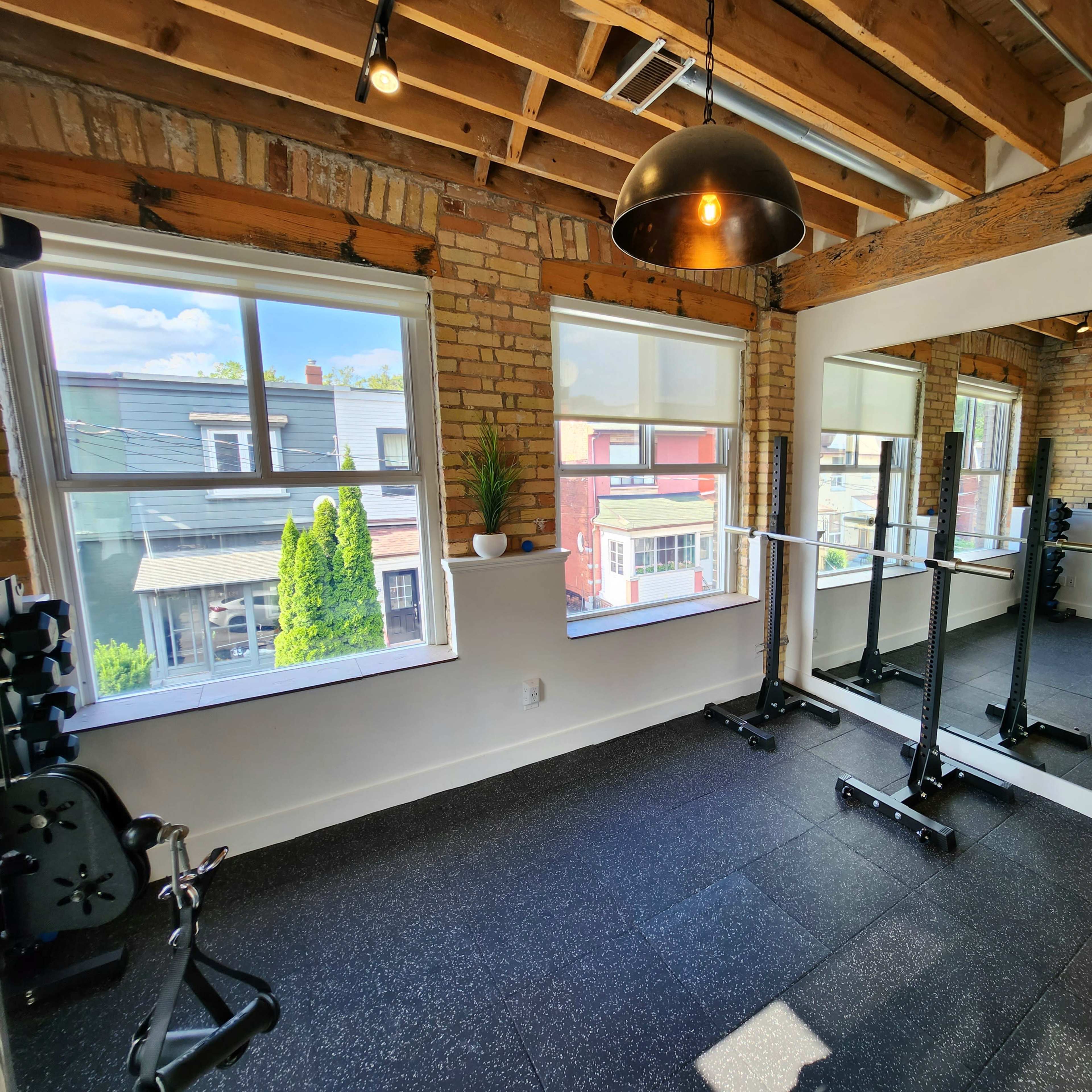 A bright gym with large windows, exposed brick walls, and a mirror, featuring weightlifting equipment and a rubber floor.