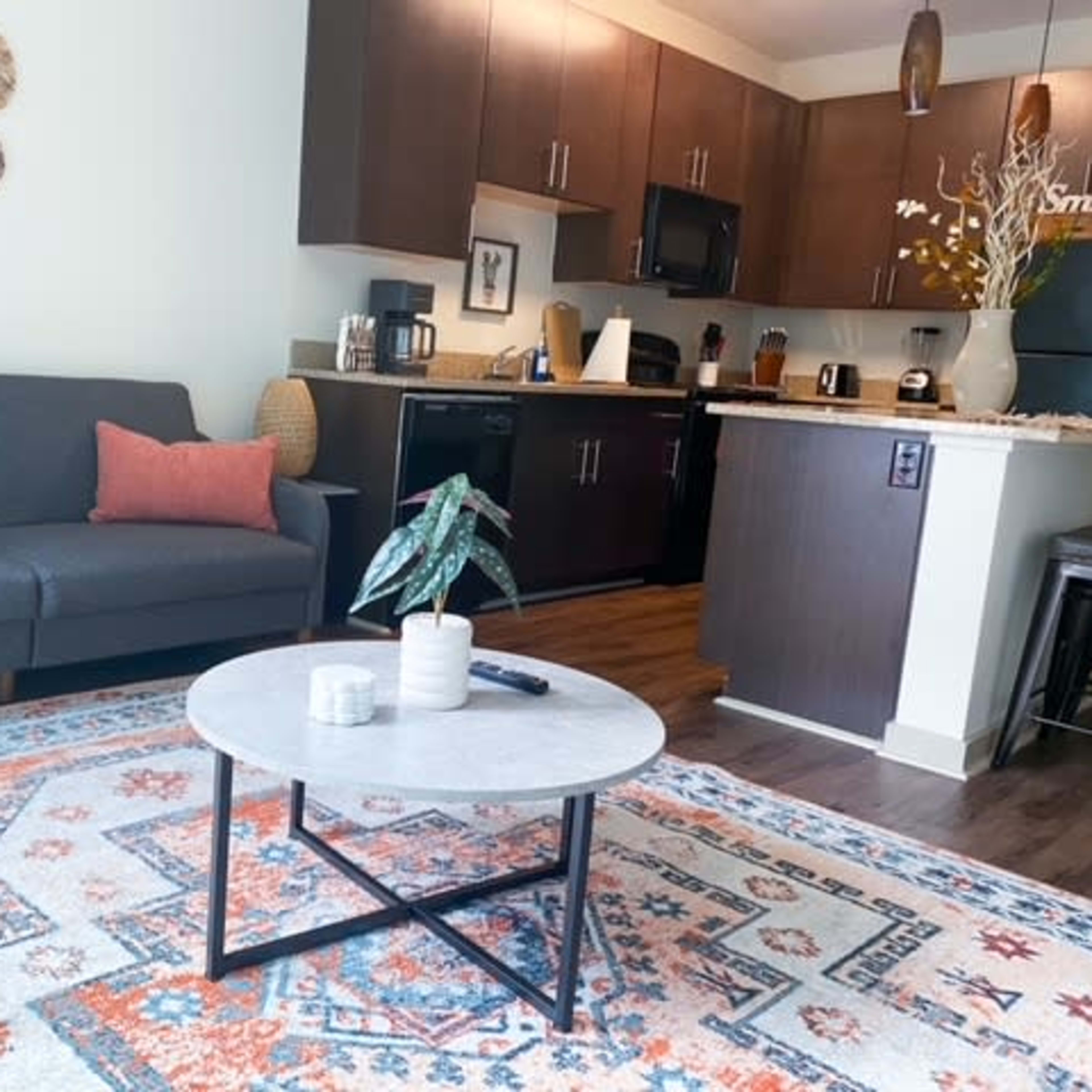 A cozy living room features a gray sofa with pink cushions, a marble-top coffee table, and a colorful area rug, adjacent to a modern kitchen with dark cabinetry.