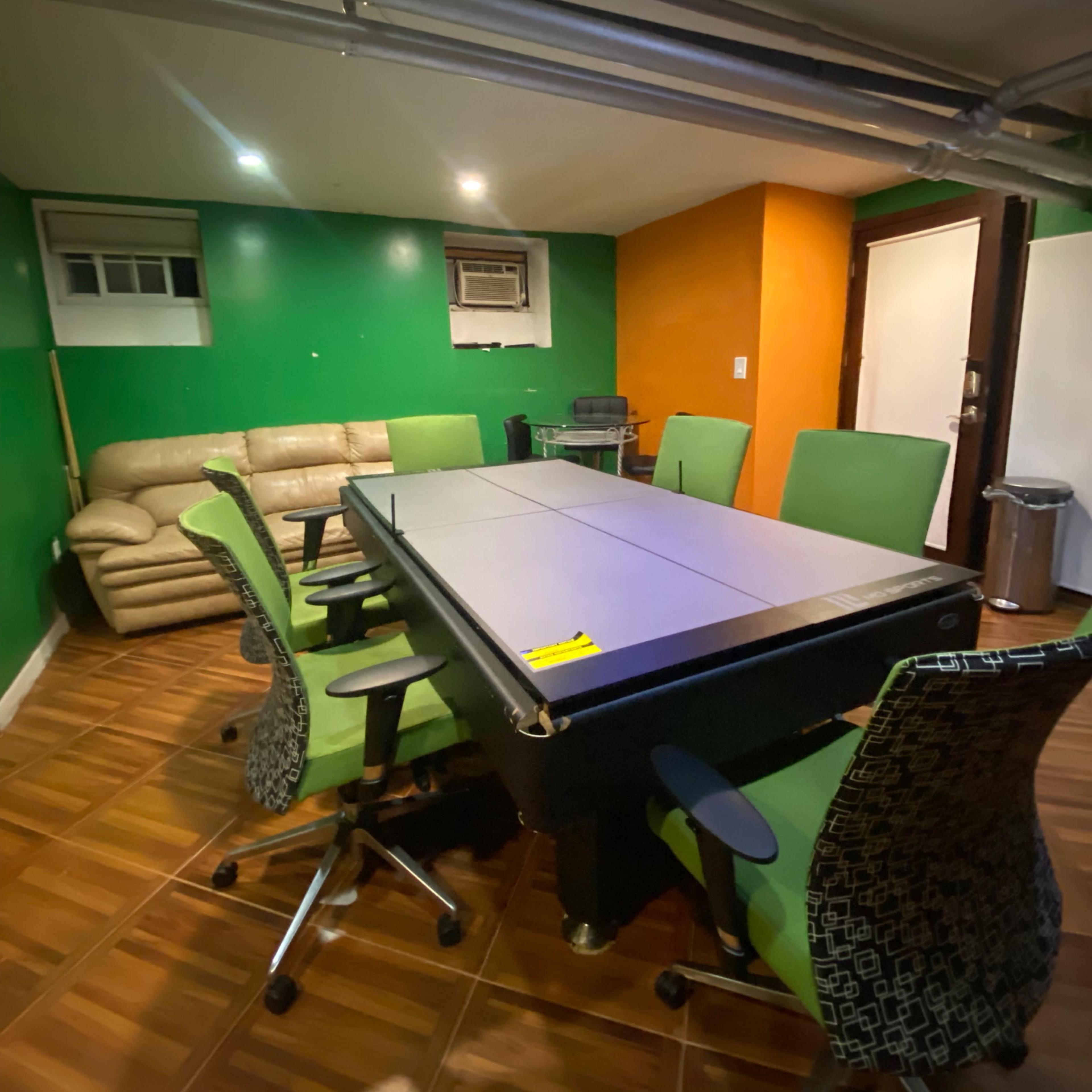 A basement room with a large table surrounded by green chairs, a beige couch in the corner, and walls painted in green and orange.