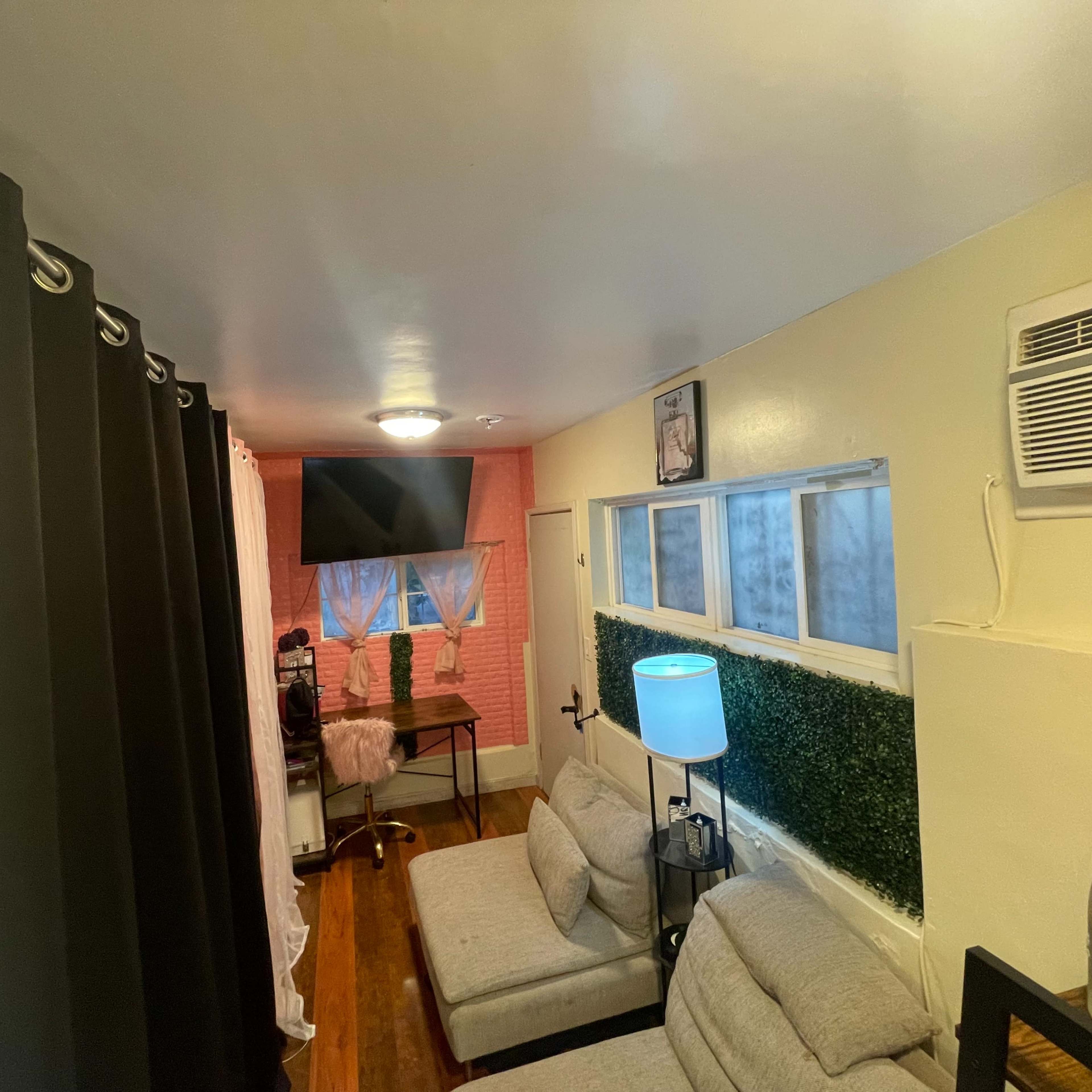 A small room with gray sofas, a desk with a chair, a television mounted on the wall, and an air conditioning unit.