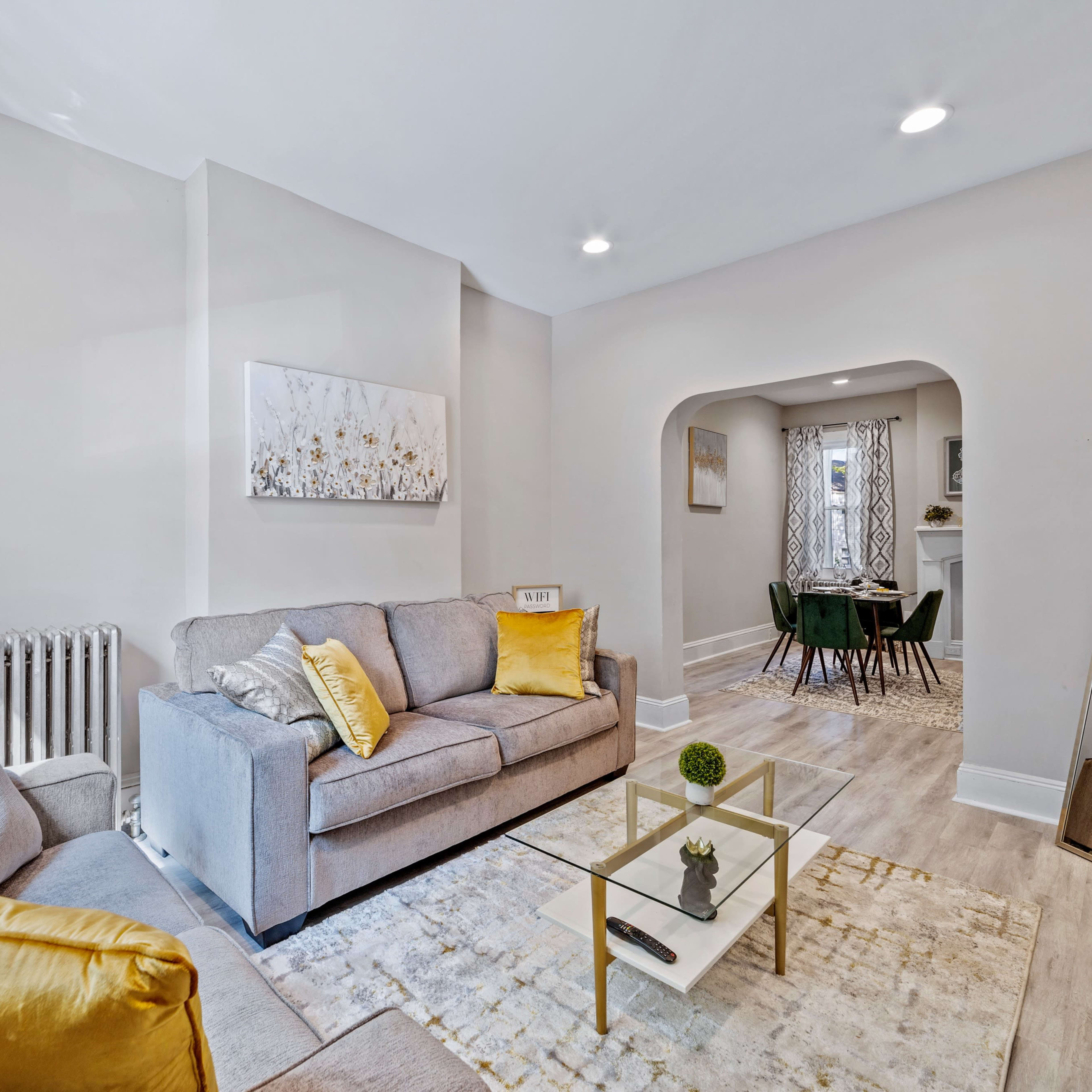 A cozy living room features a gray couch with yellow accents, a glass coffee table on a light rug, and an archway leading to a dining area.