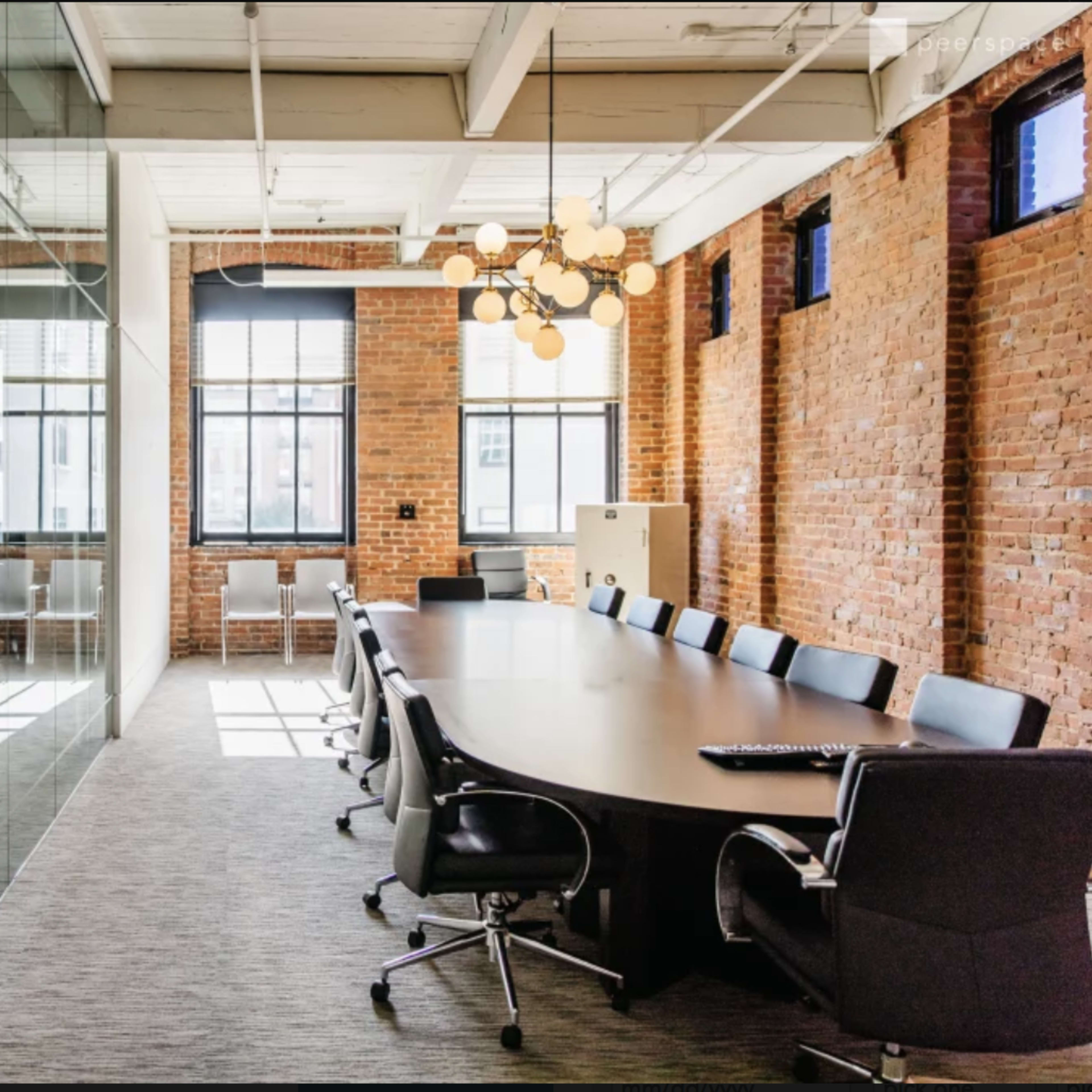 A modern conference room with a large oval wooden table surrounded by black chairs, brick walls, and large windows allowing natural light.