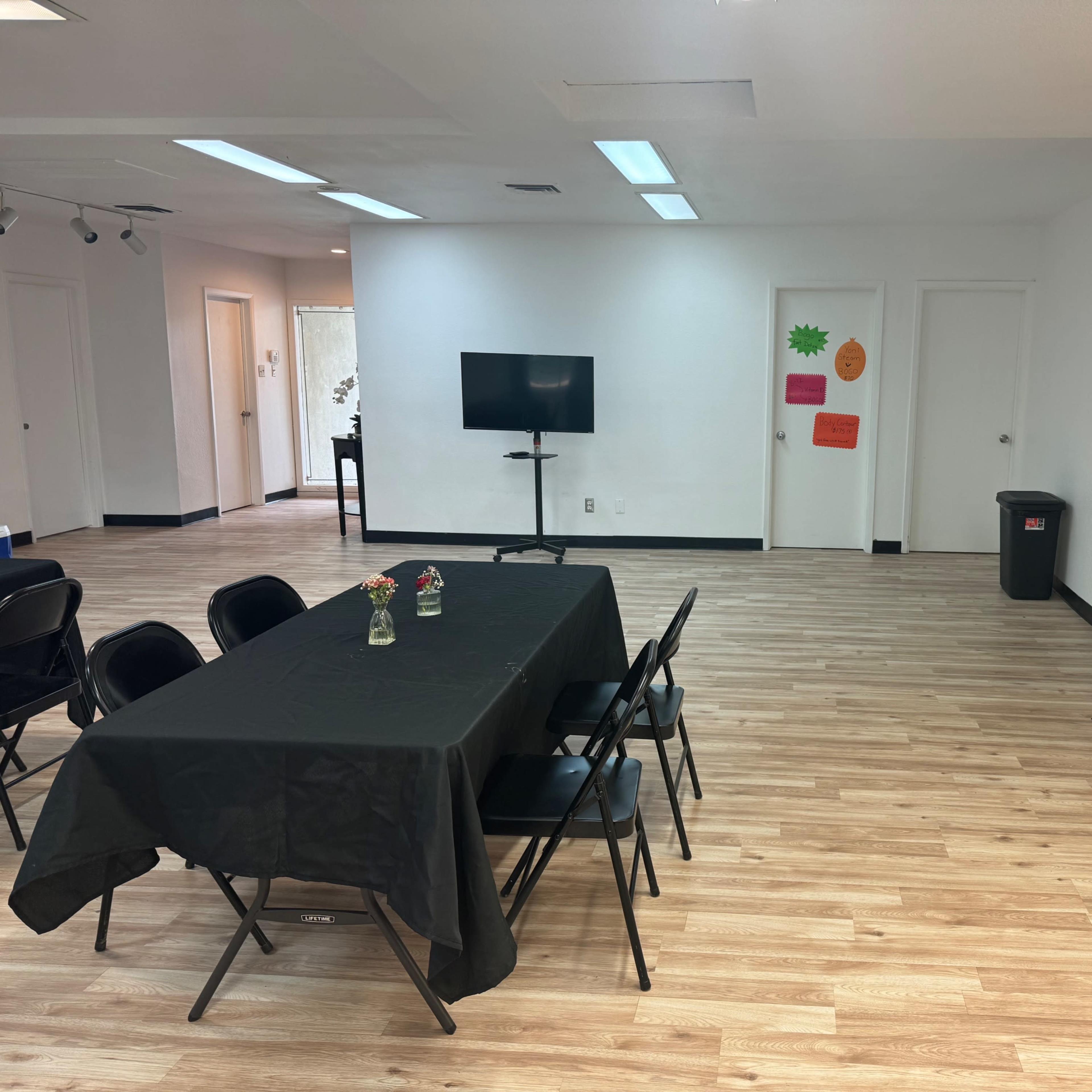 A spacious room with light wood flooring, featuring two tables covered with black tablecloths and several chairs arranged nearby, along with a television mounted on a stand and a trash bin.