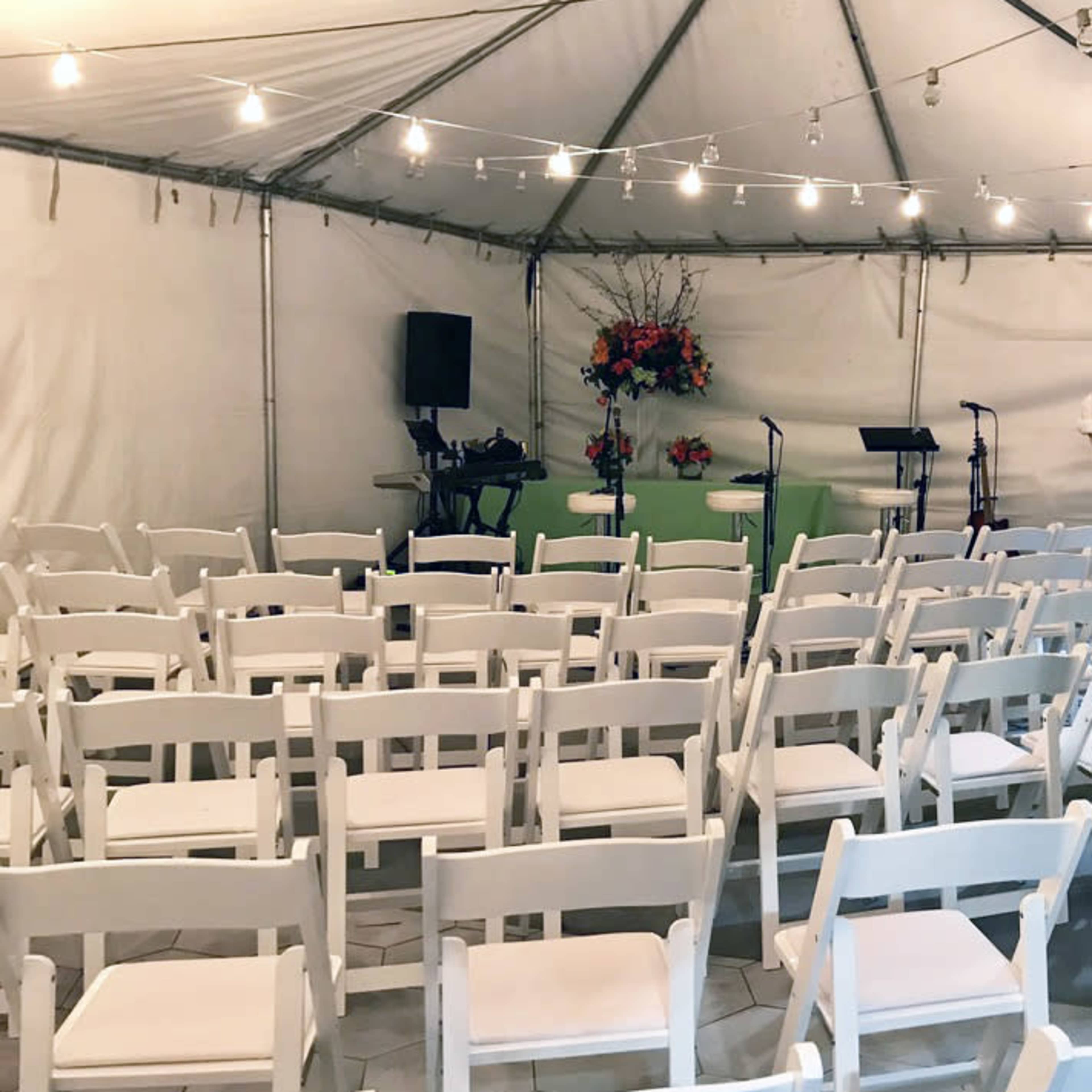 A tented space with white chairs arranged in rows facing a small stage decorated with flowers and musical equipment.