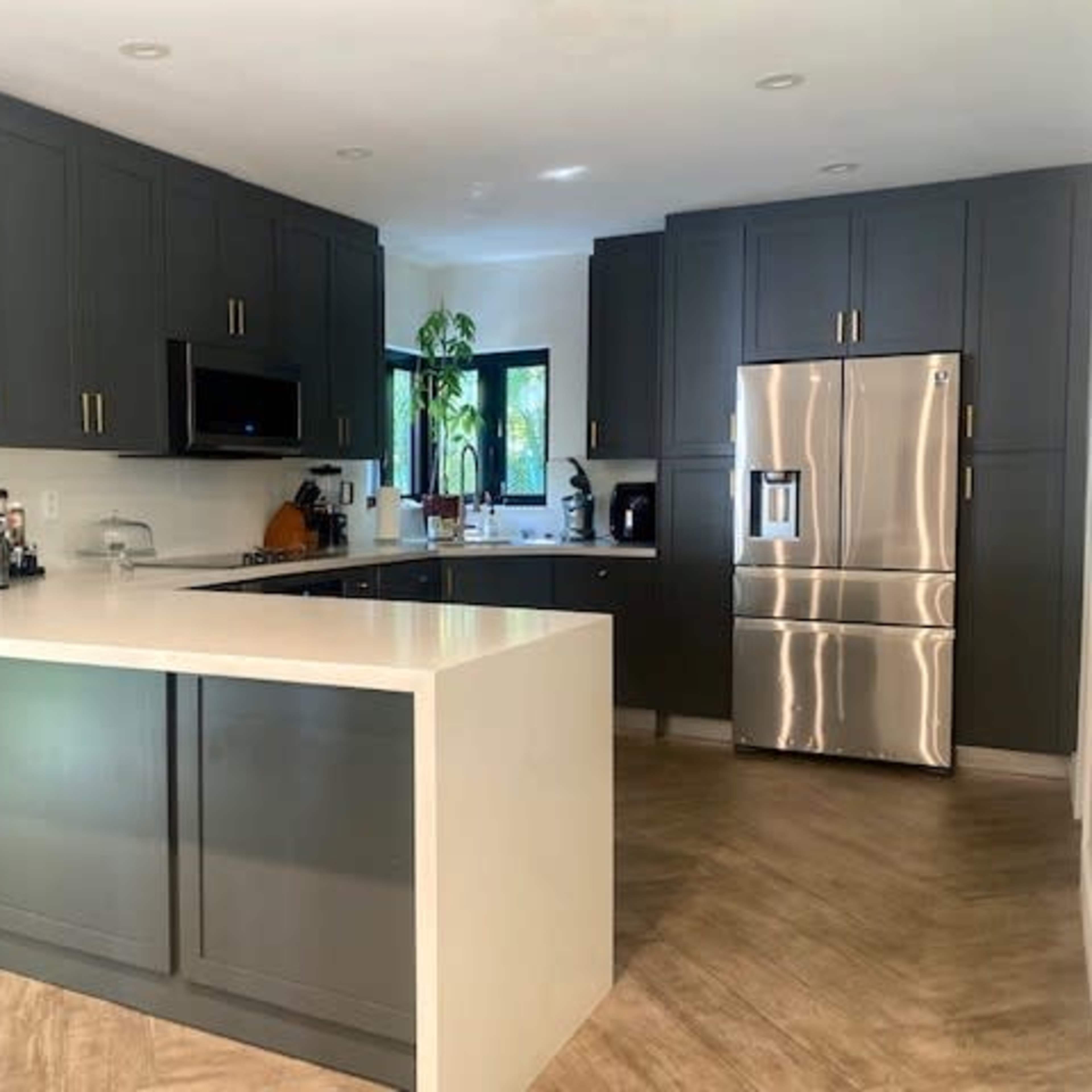 The kitchen features dark cabinets, a stainless steel refrigerator, and a large white countertop with a view of a window.