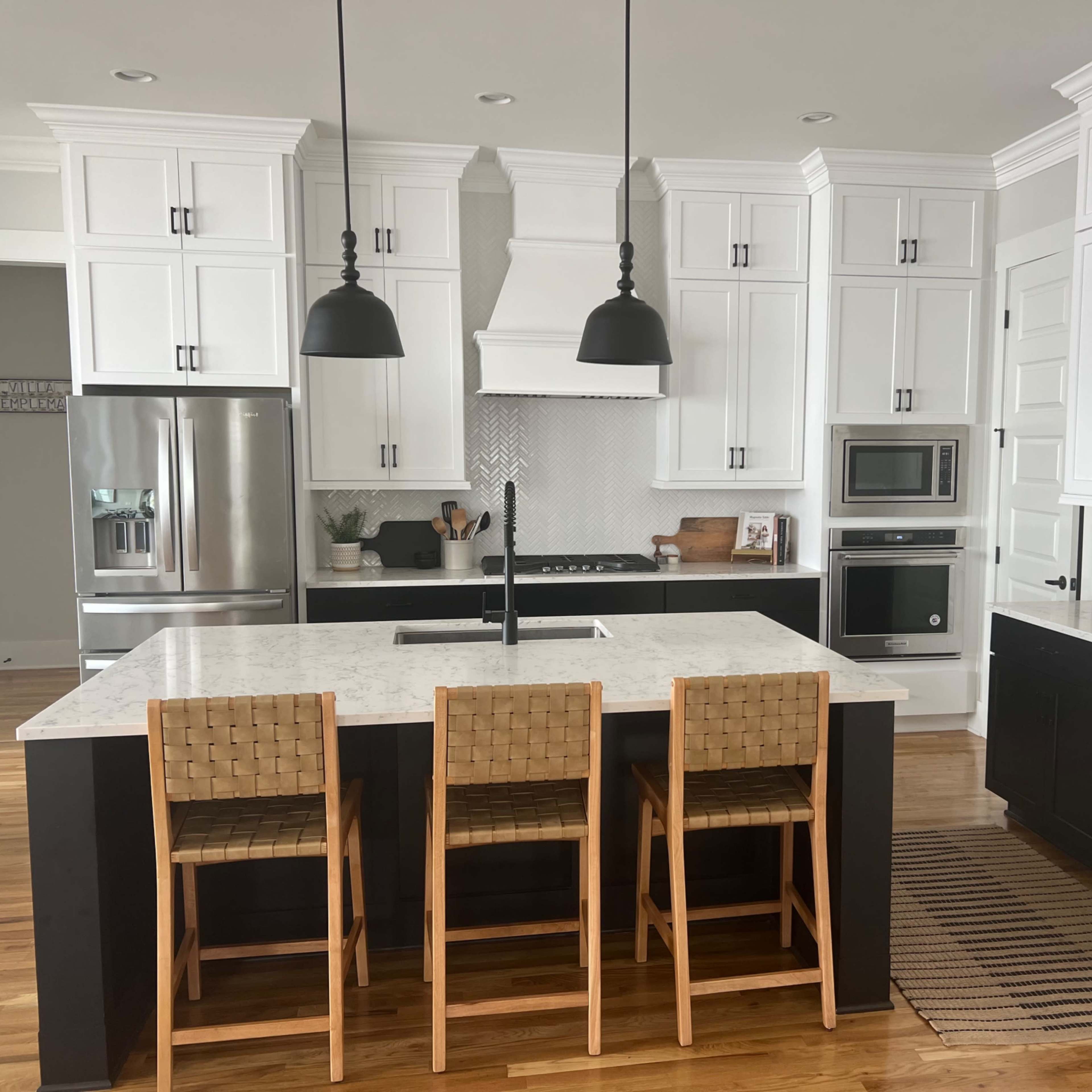 A modern kitchen with white cabinetry, a central island with three wooden stools, stainless steel appliances, and pendant lighting.
